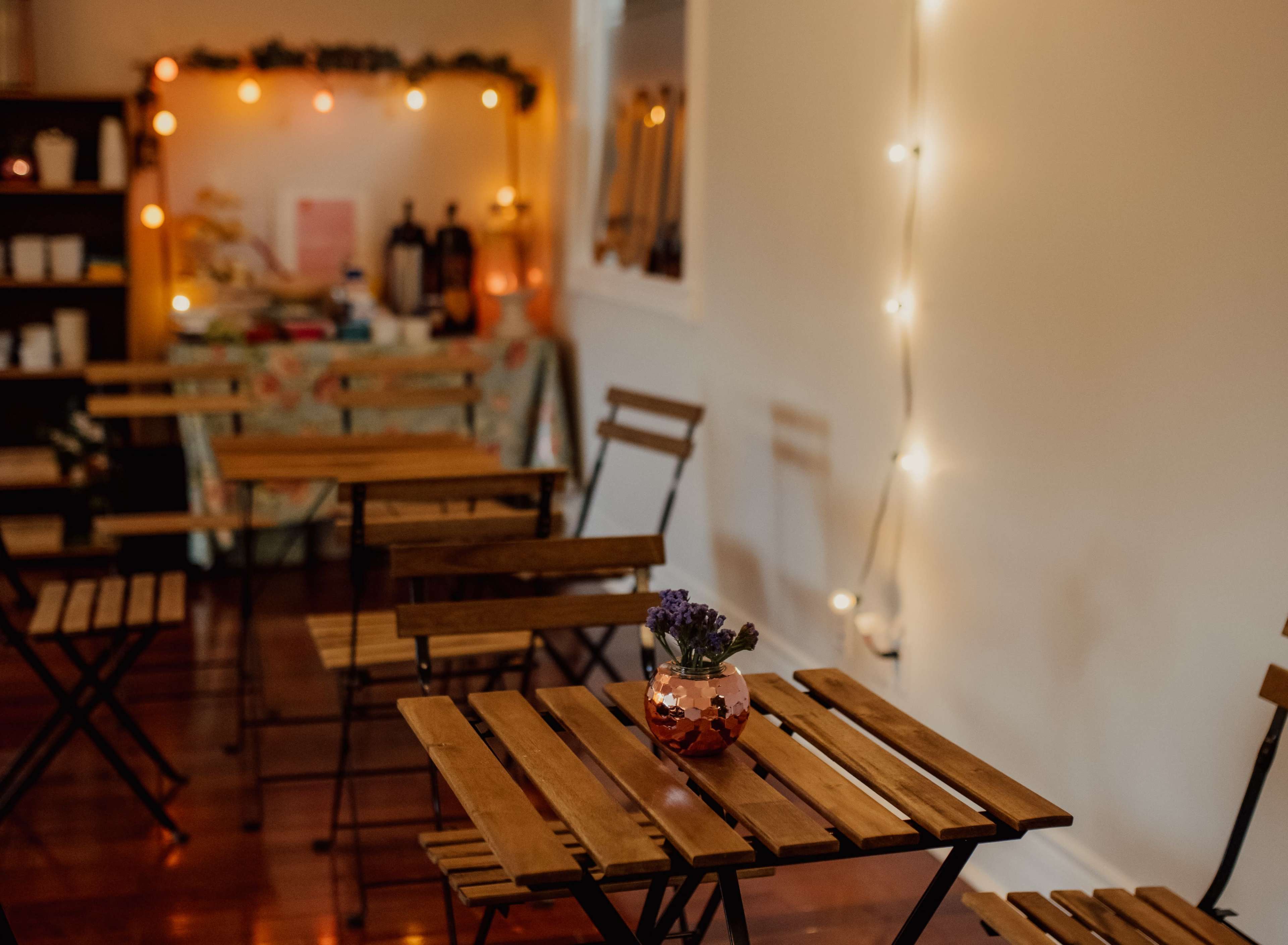 A cozy room features wooden folding tables with a small glass vase of lavender at the center, illuminated by warm string lights.