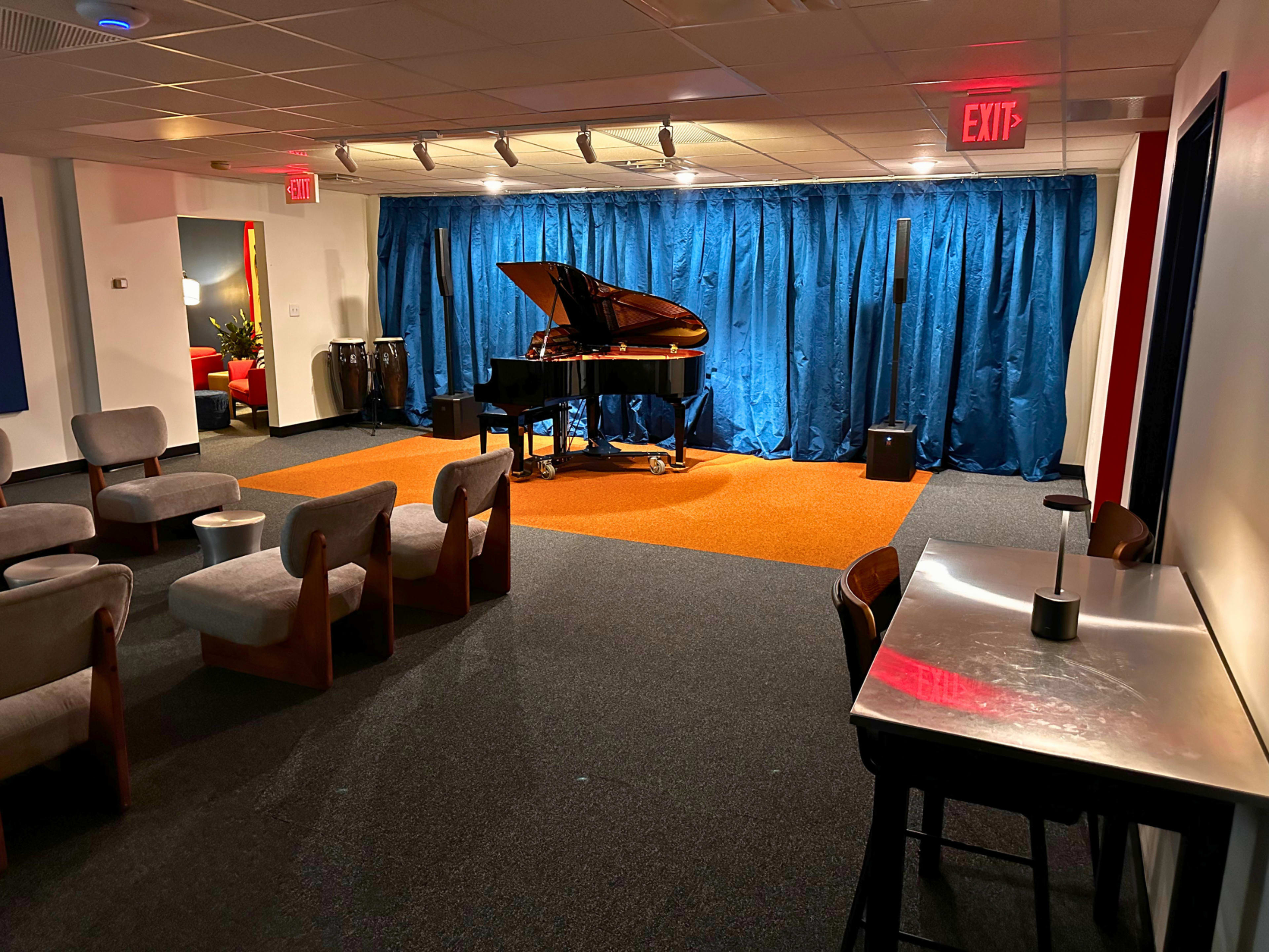 The image shows a cozy room with a grand piano on a stage, surrounded by blue curtains, with chairs and tables arranged in the foreground.