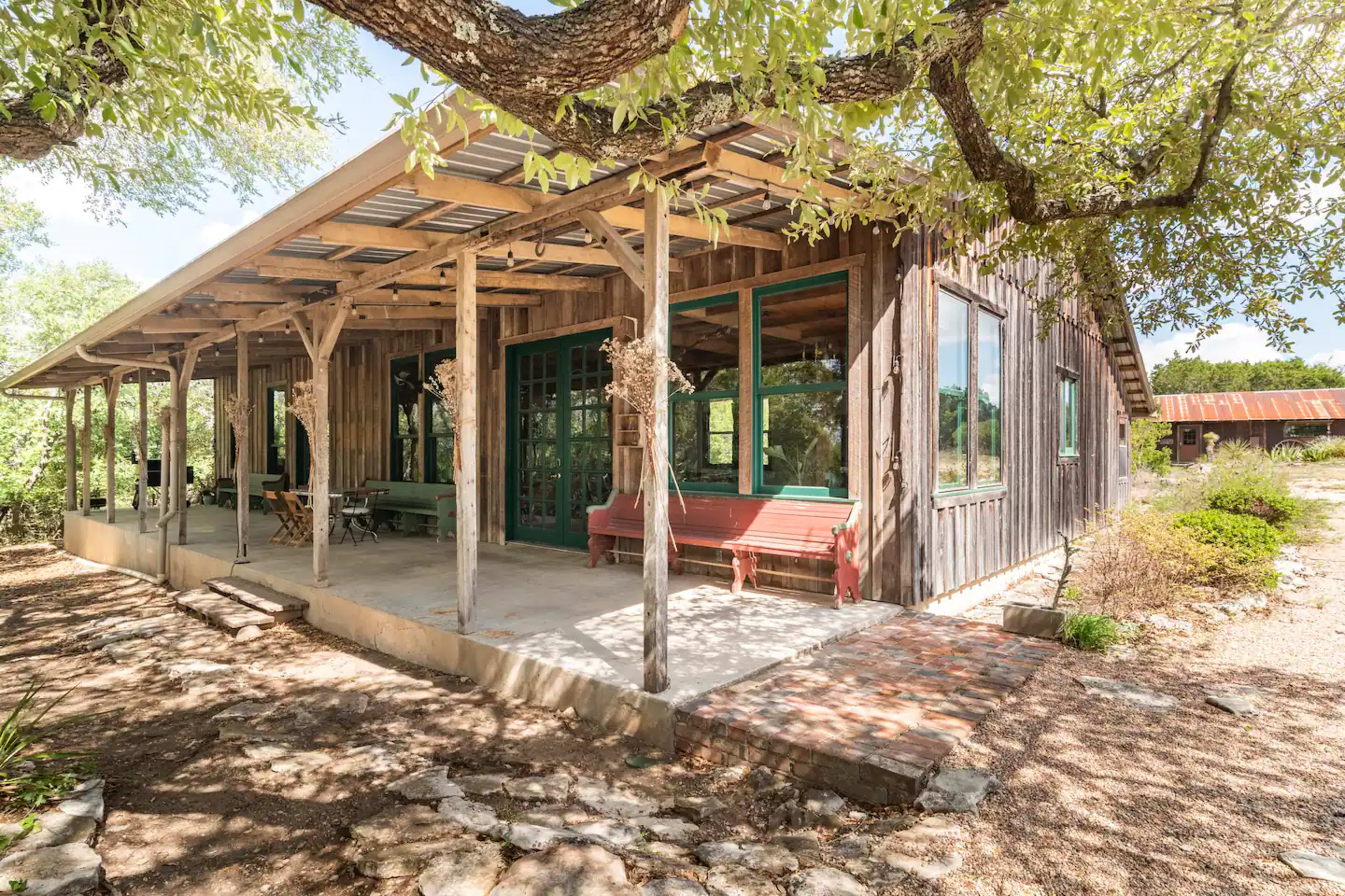 The image shows a rustic wooden house with a spacious porch, surrounded by greenery and a gravel pathway.