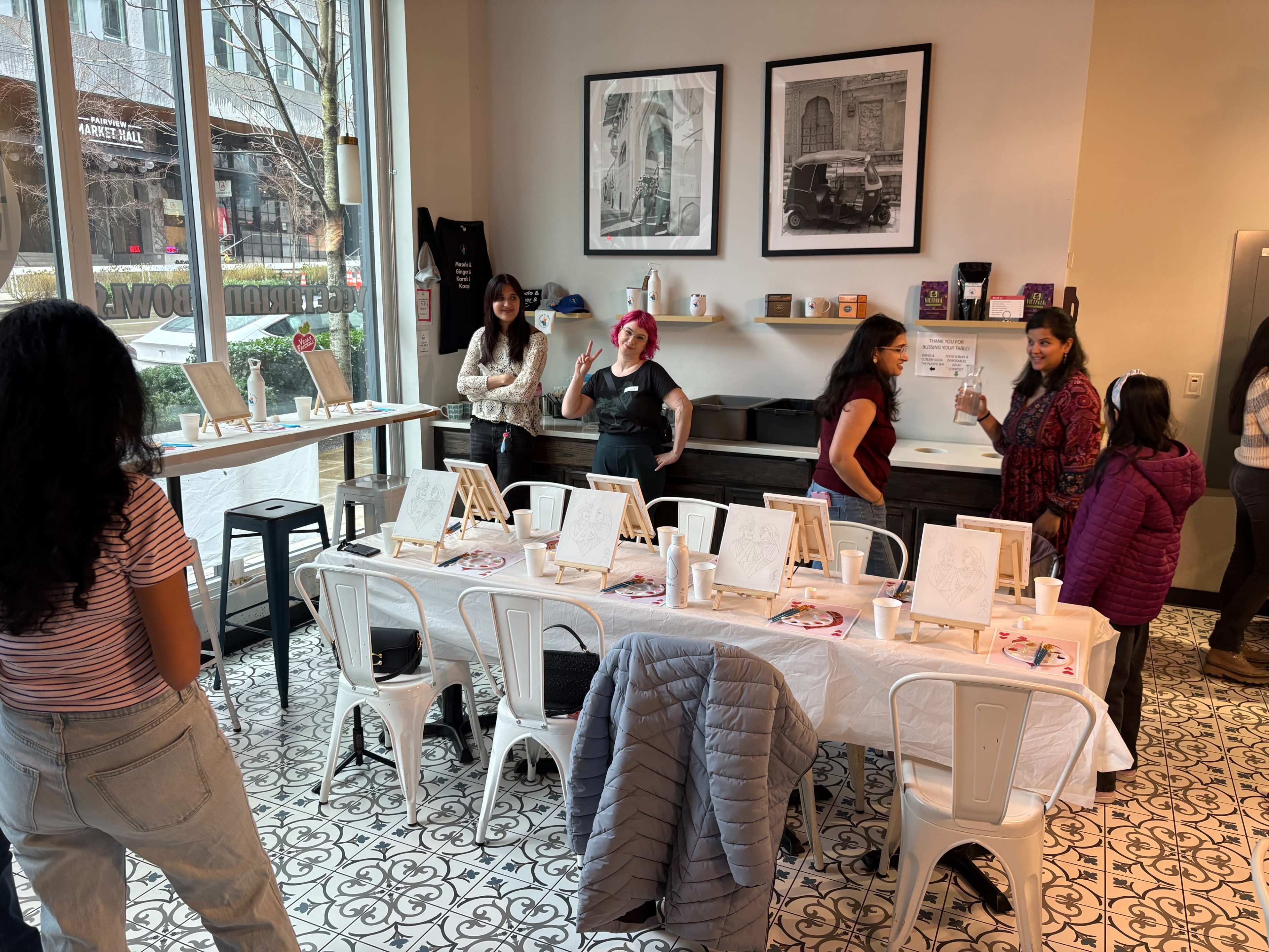 A group of people are preparing for a painting class in a well-lit café, with easels and art supplies set up on a long table.