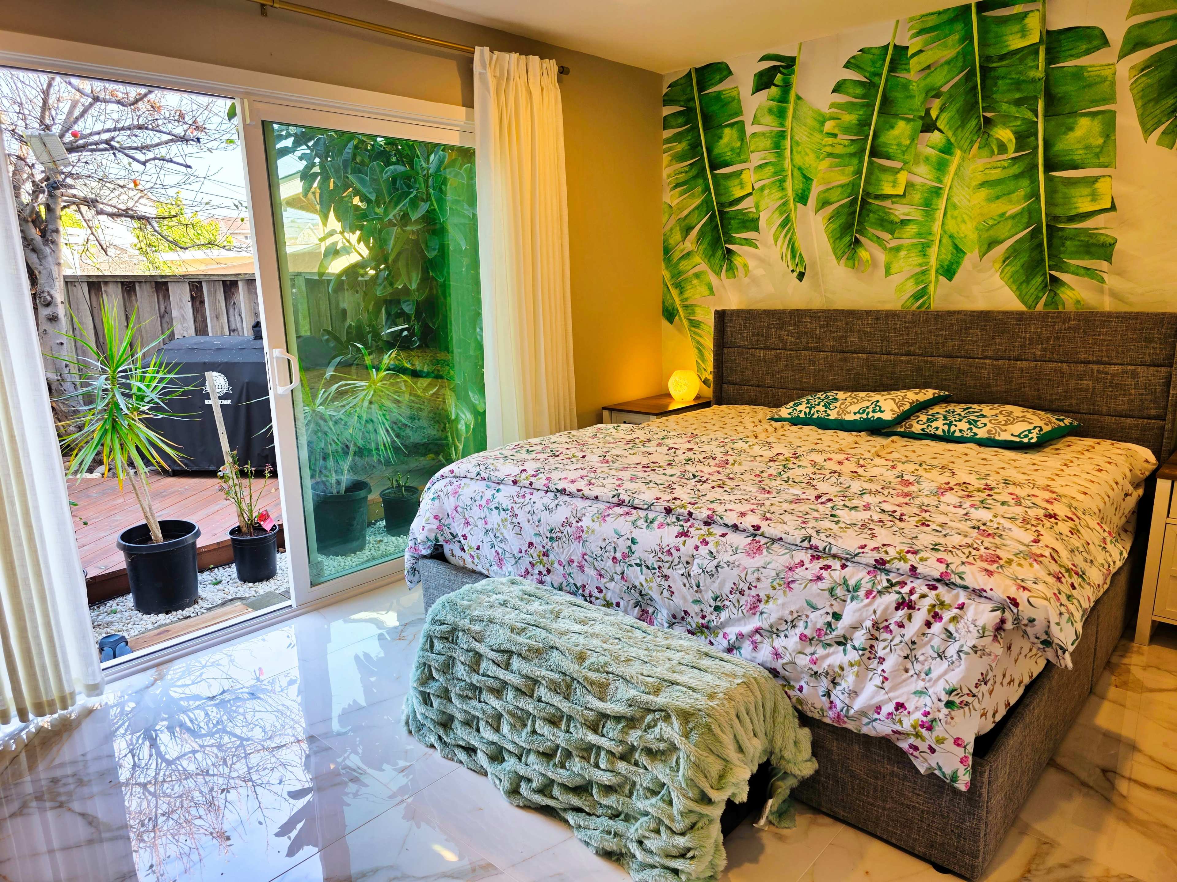 The image shows a brightly lit bedroom with a large bed adorned with a floral-patterned blanket, a wall decorated with green leaf murals, and a sliding glass door leading to an outdoor area.