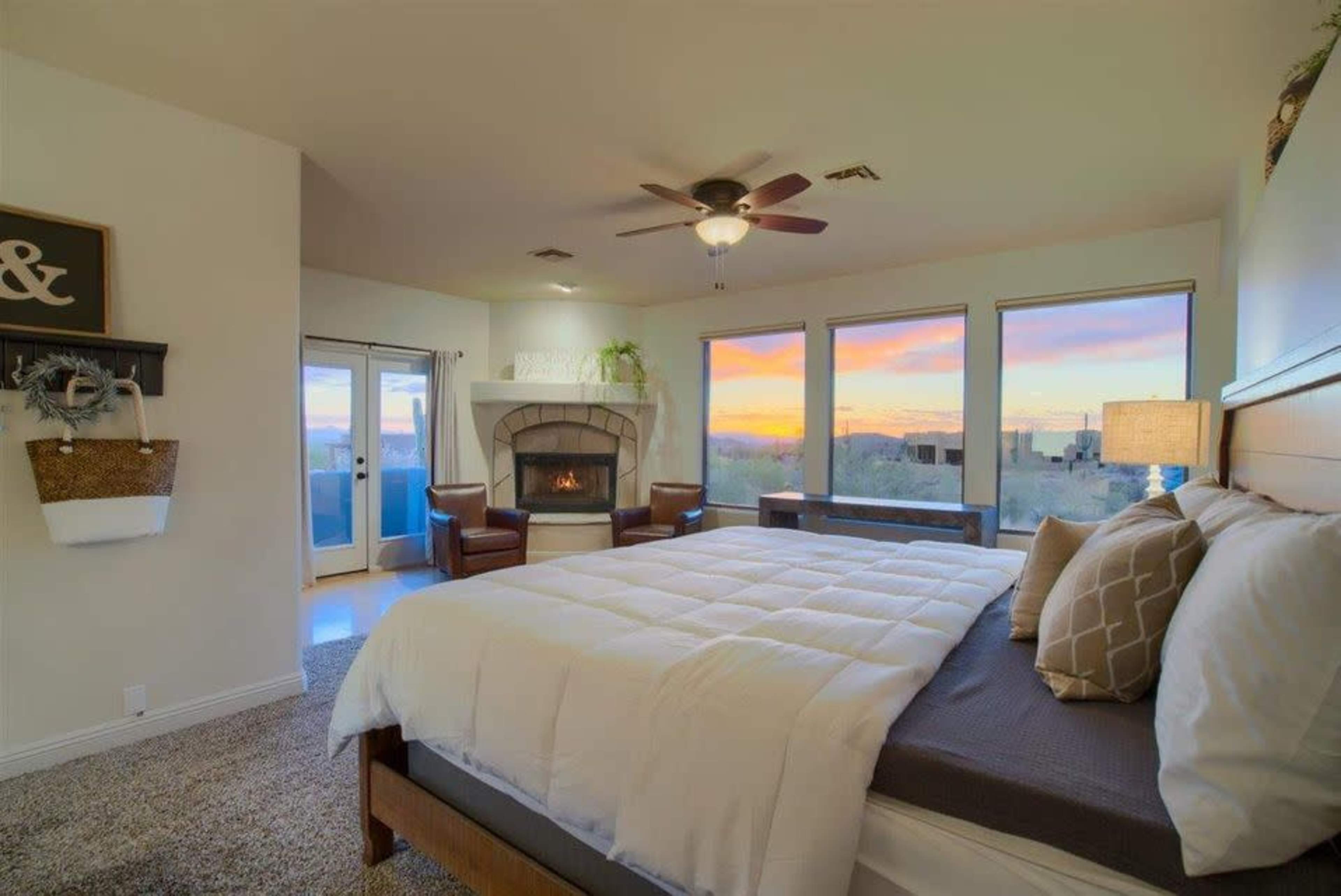 A spacious bedroom features a large bed with a white comforter, a fireplace, and three windows displaying a colorful sunset.