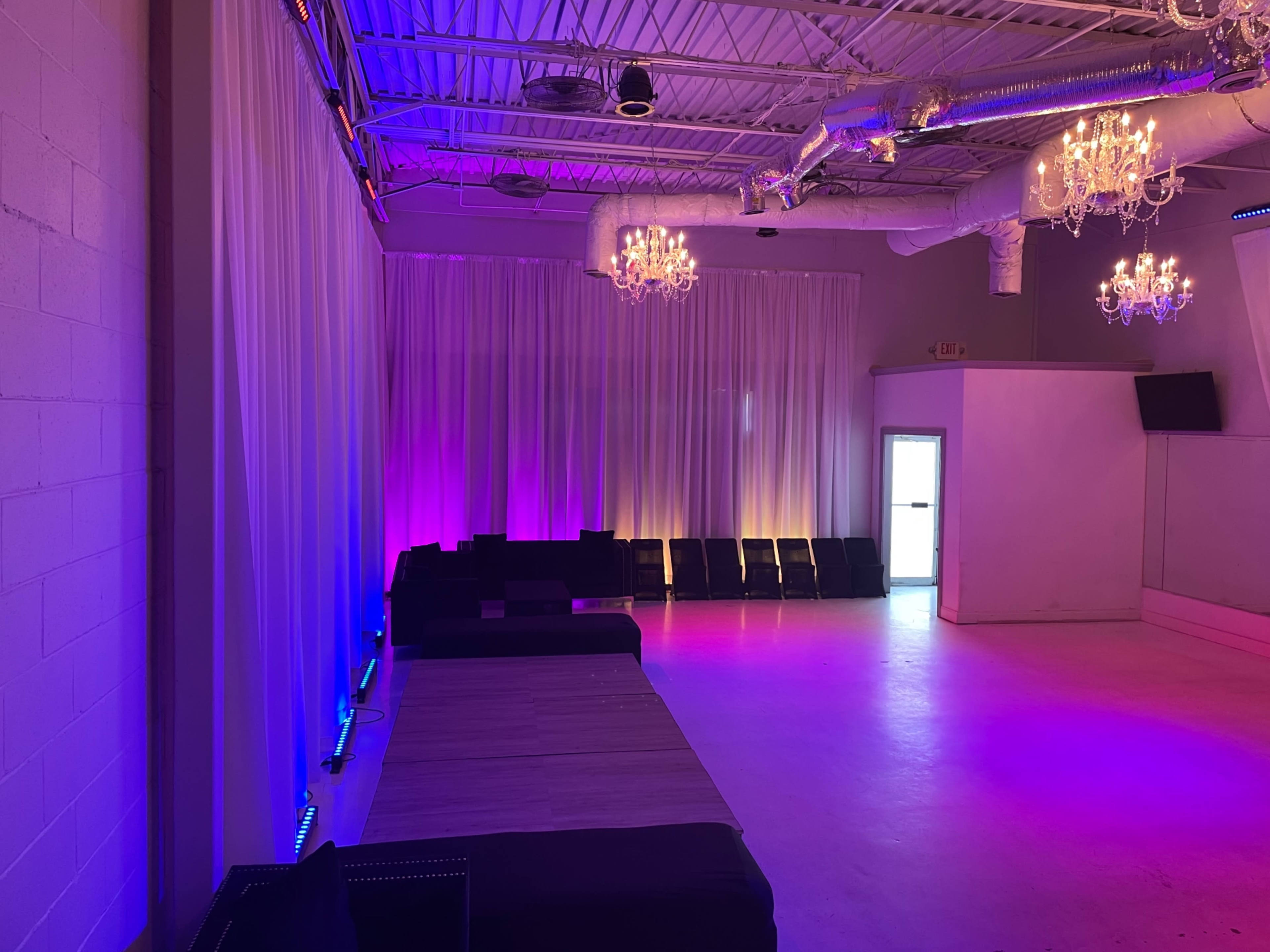 The image shows an empty event space with purple and blue lighting, featuring white drapes and several chandeliers.