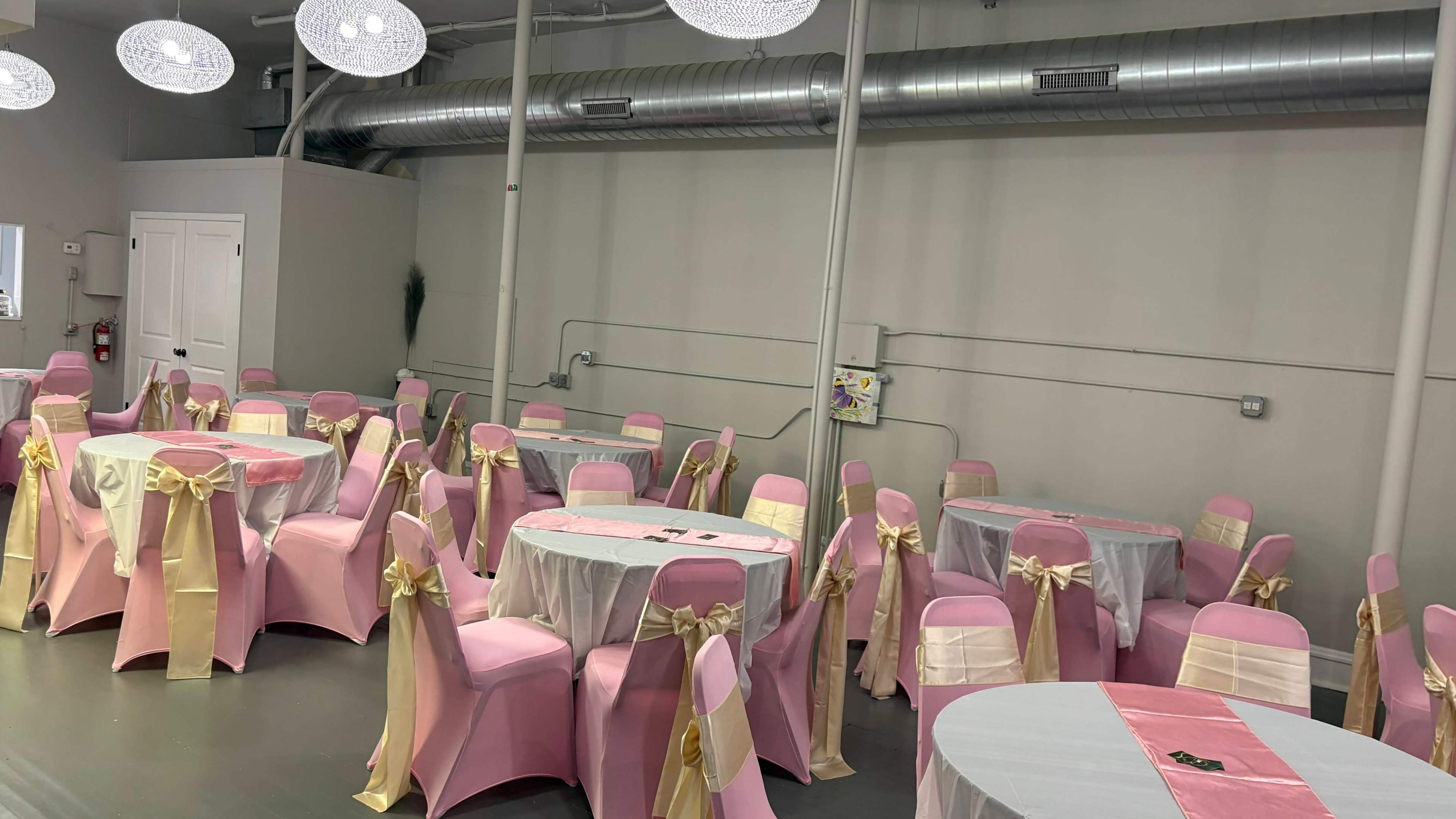 The image shows a banquet room set up with round tables covered in white and pink tablecloths, surrounded by pink chairs adorned with gold sashes.