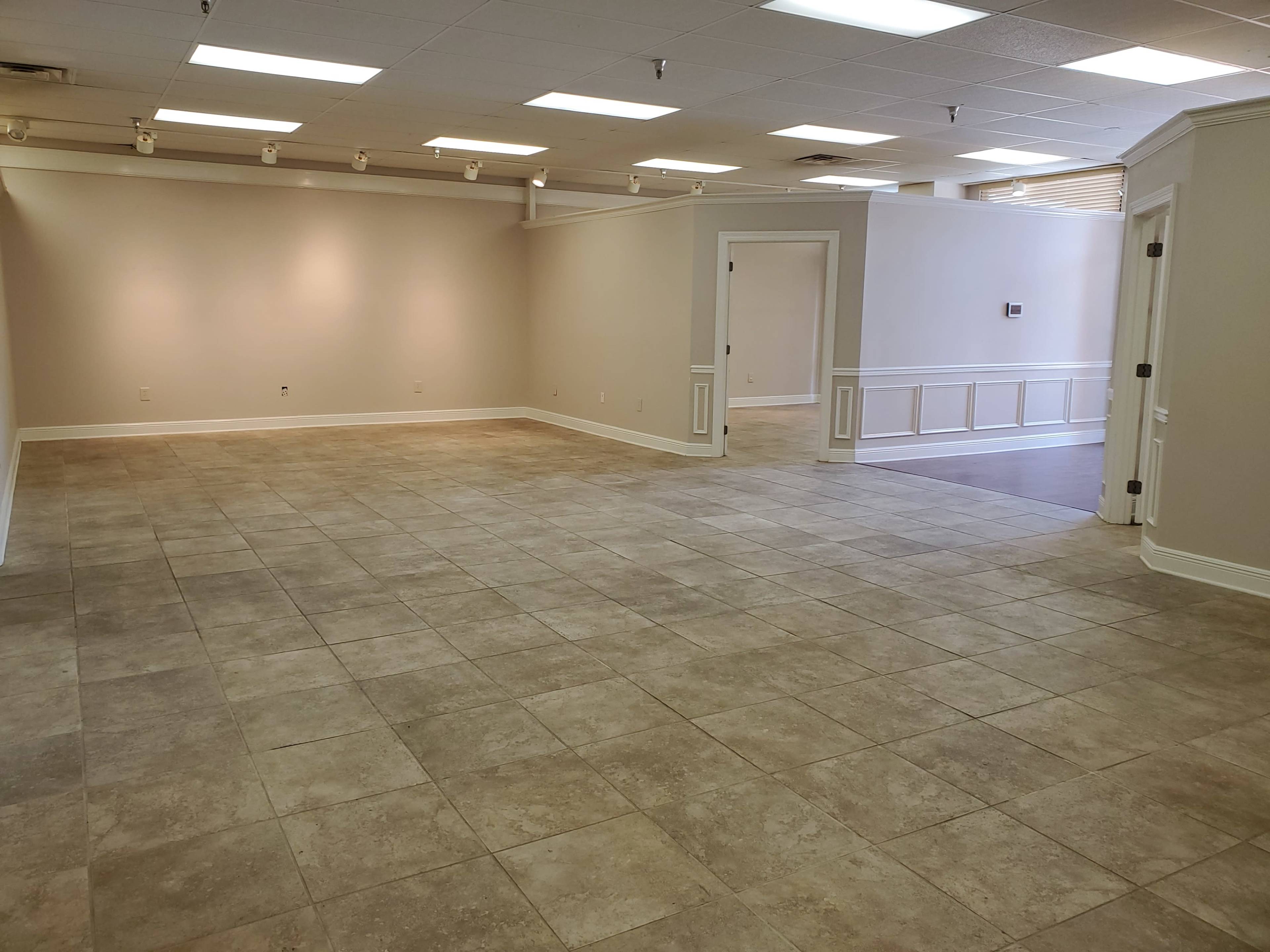 The image depicts a spacious, empty room with tiled flooring and light-colored walls, featuring doorways leading to adjacent areas.