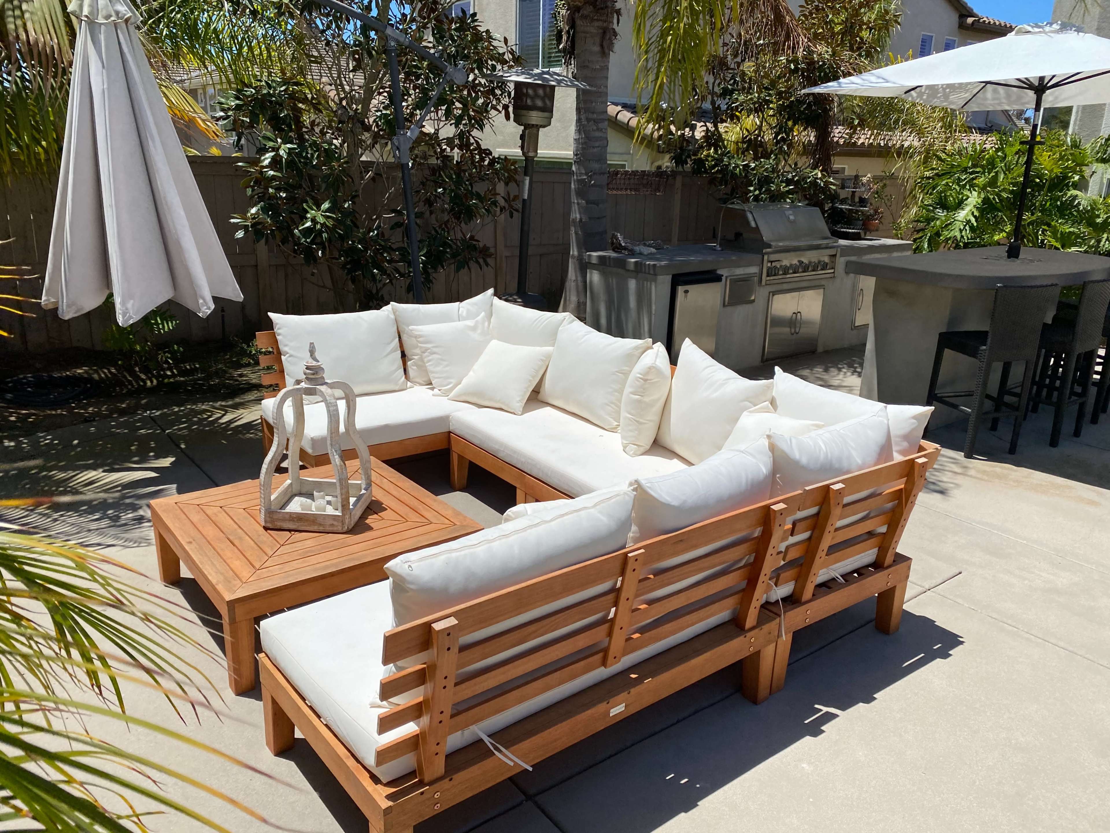 A wooden outdoor seating area with white cushions, a lantern on a coffee table, and an umbrella nearby.