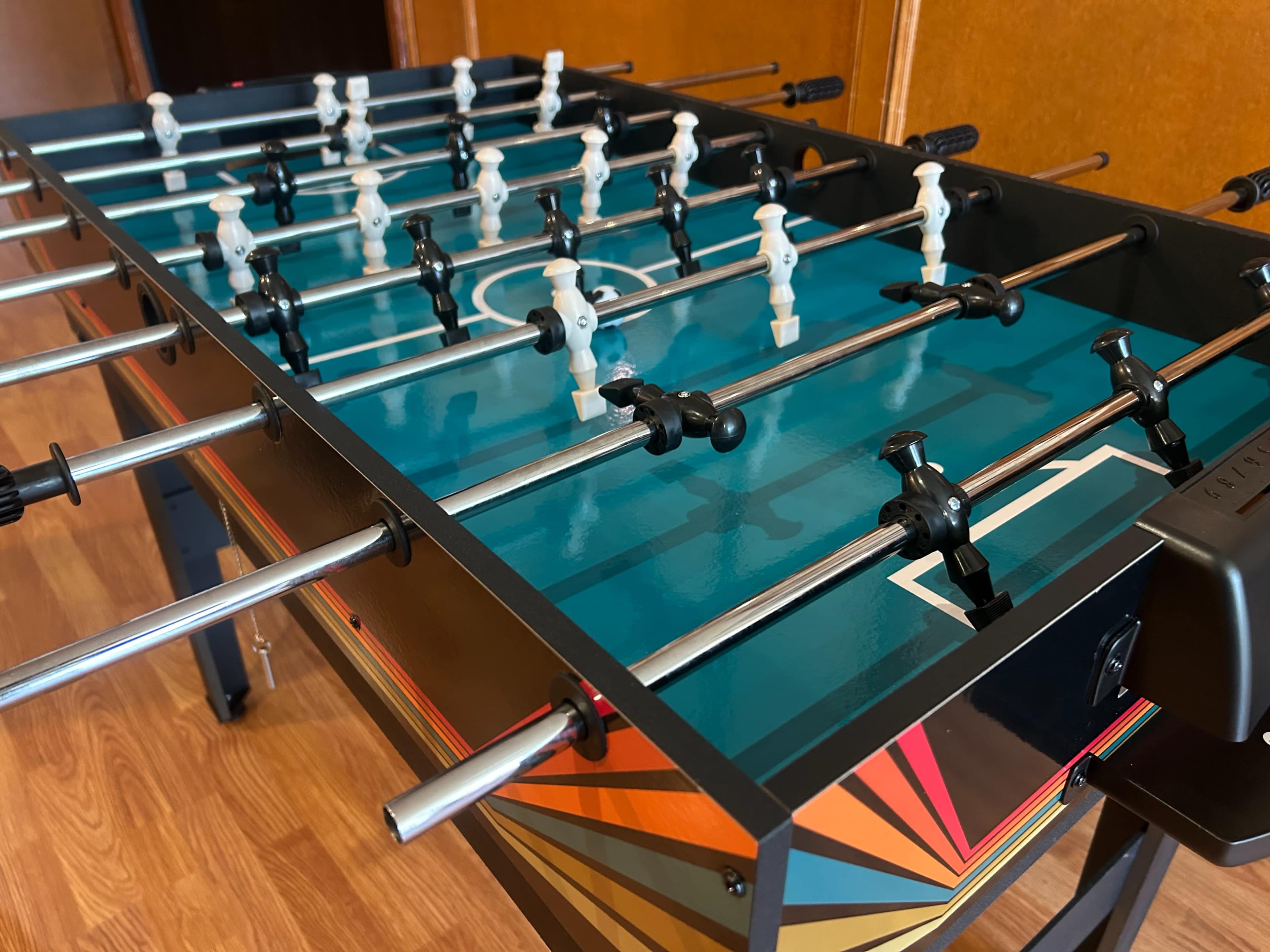 The image shows a foosball table with a colorful design, featuring rows of player figures on rods and a game control panel on the side.