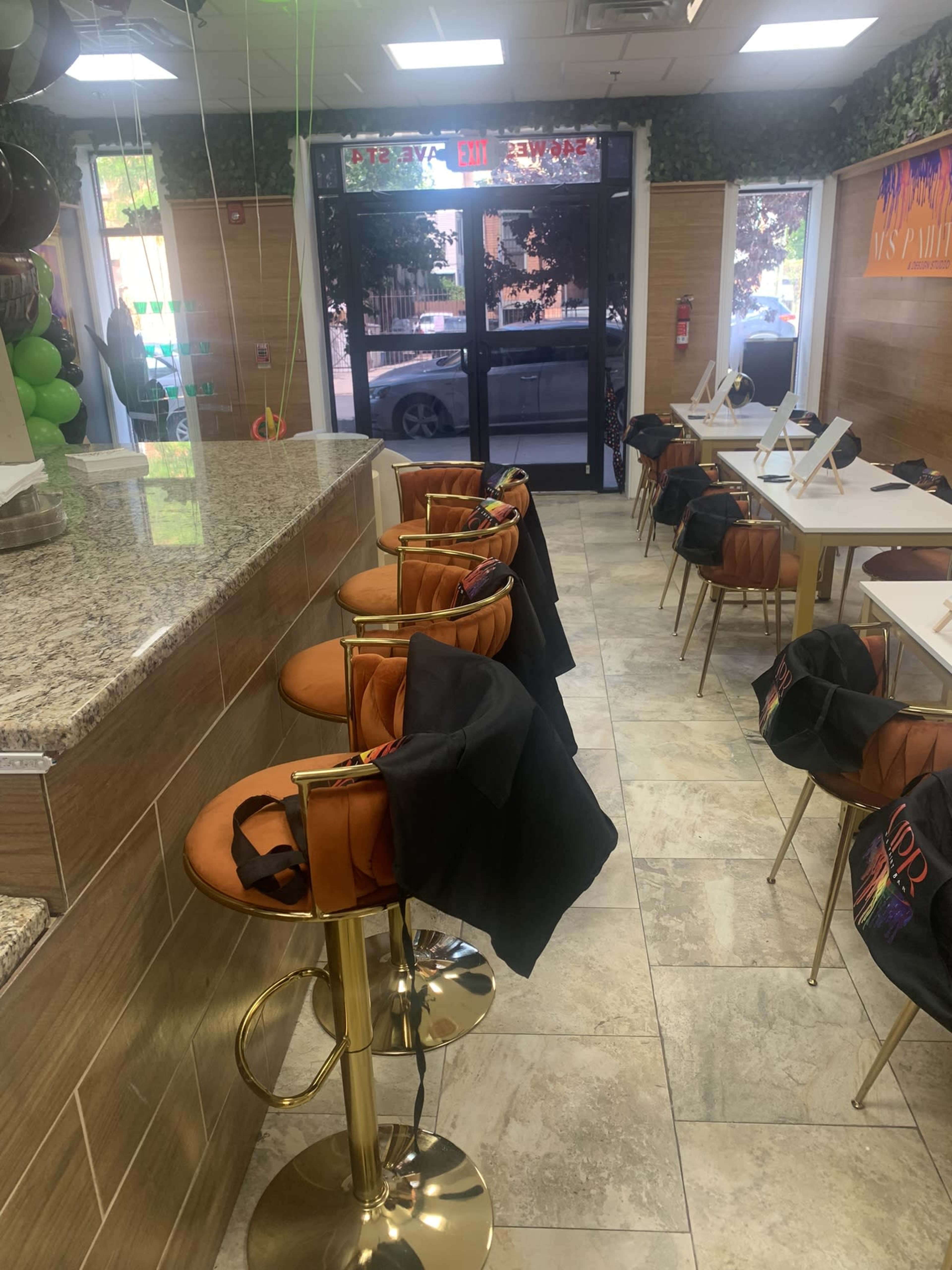 The image shows a café interior with several empty golden stools at a counter and tables lined with black bags.