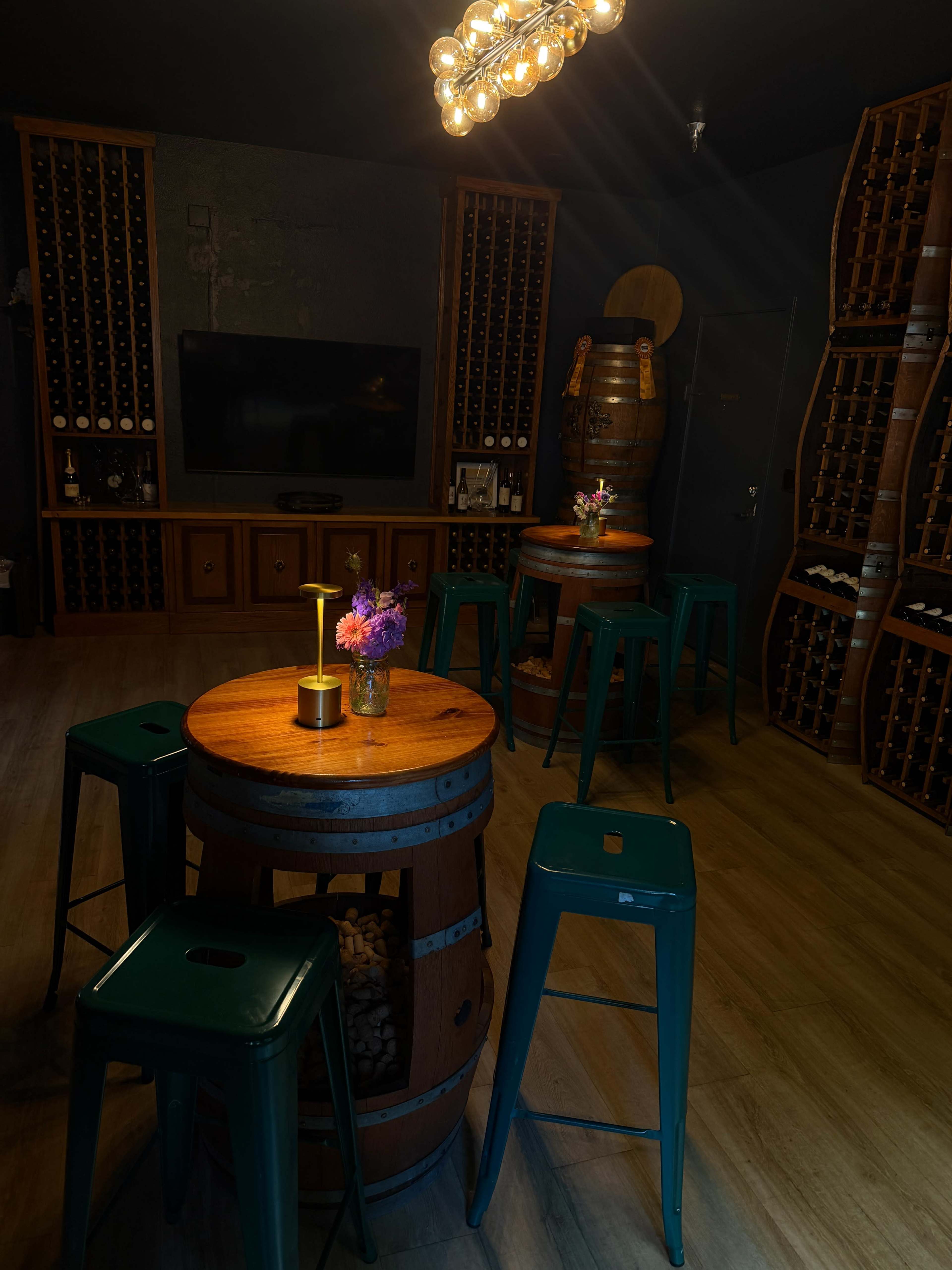 The image shows a dimly lit wine tasting room with wooden barrel tables, green metal stools, and wine bottle storage against the walls.