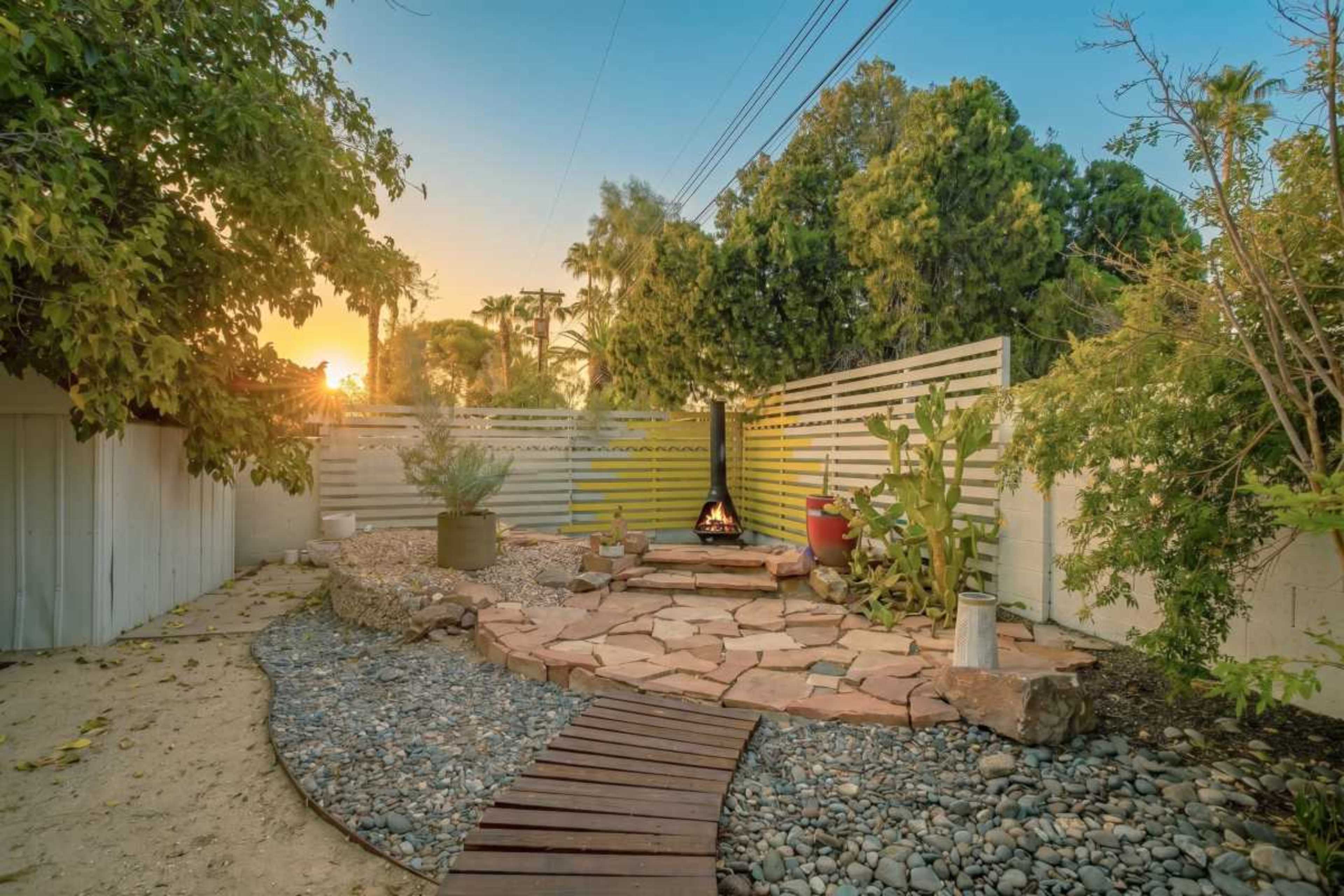 The image shows a small backyard with a stone pathway leading to a fire pit surrounded by gravel and lush greenery, with a sunset in the background.
