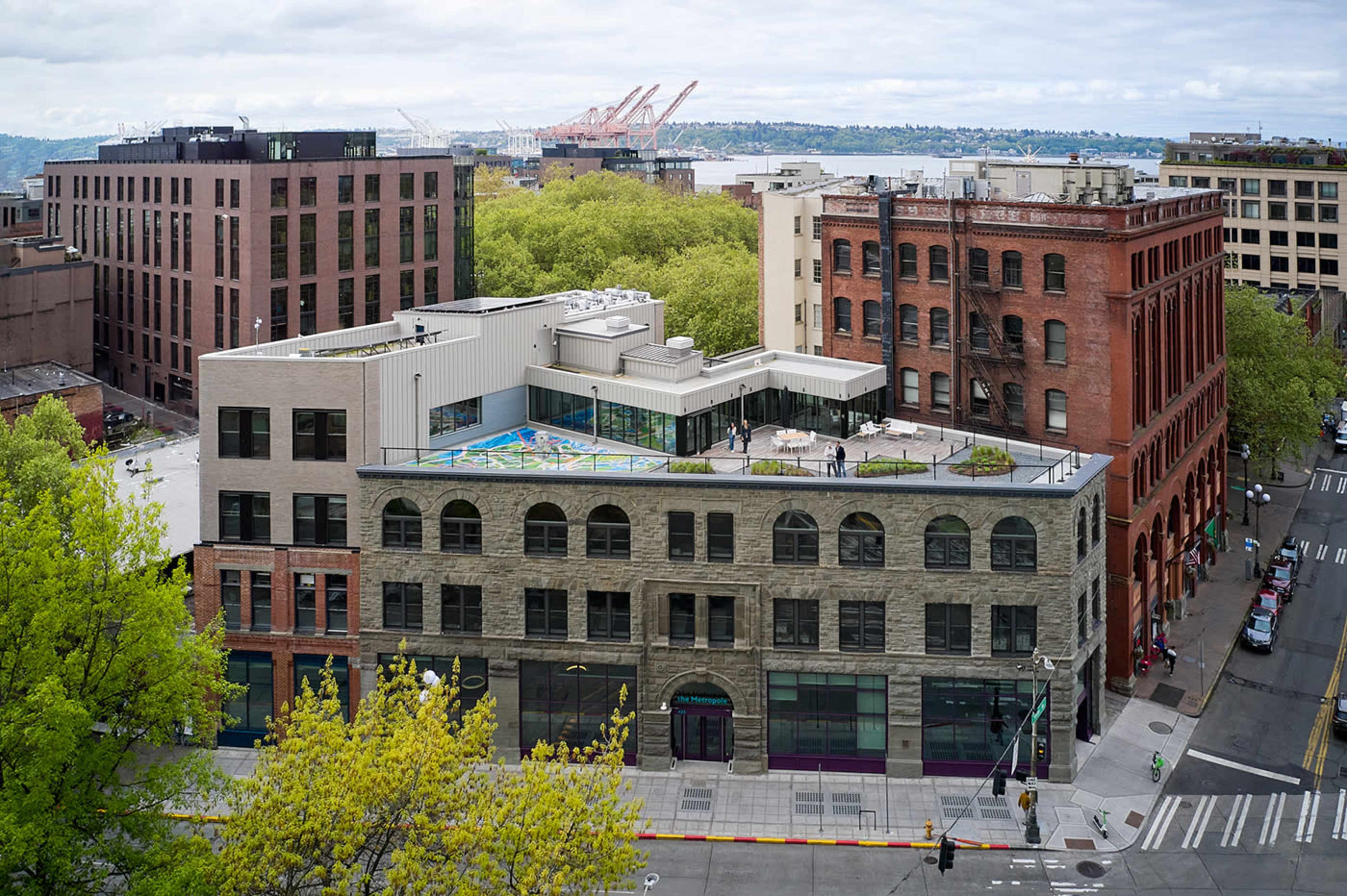 The image shows a multi-story building featuring a flat rooftop with a colorful design, surrounded by trees and other buildings in an urban setting.