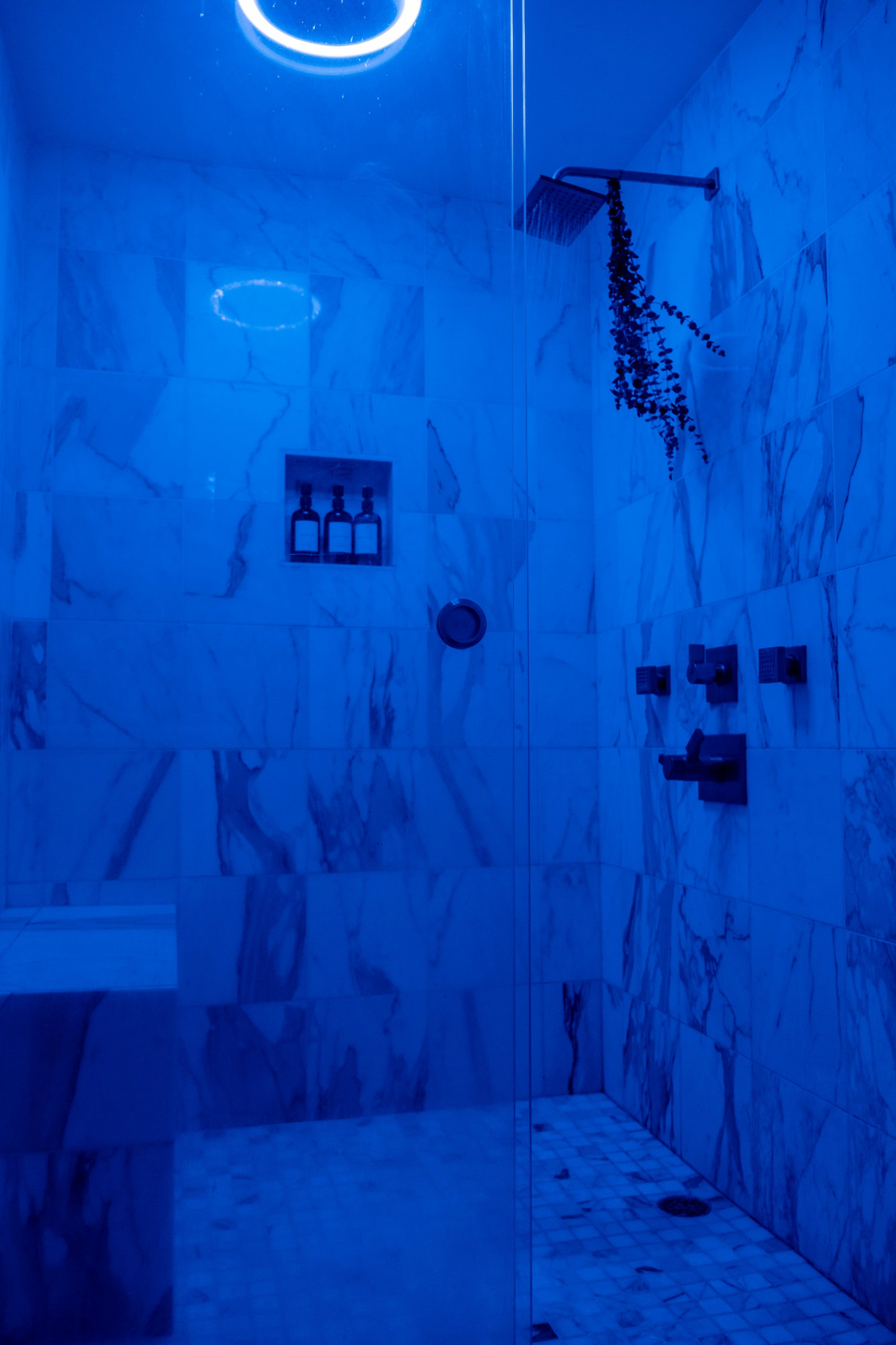 A modern shower with marble walls, illuminated by blue lighting and featuring a wall-mounted showerhead and shelf with bottles.
