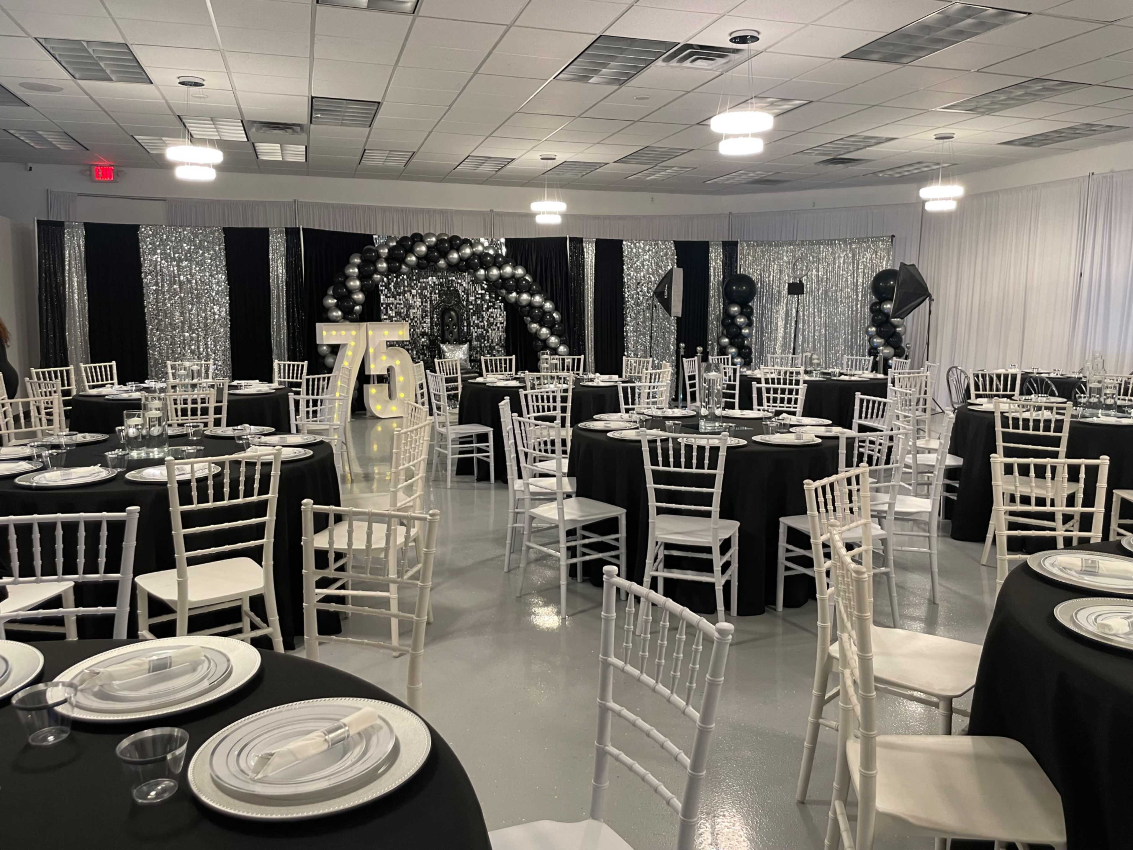 The image shows a decorated banquet hall set for a event, featuring black and white tables, a large "75" sign, and balloon arch decorations.