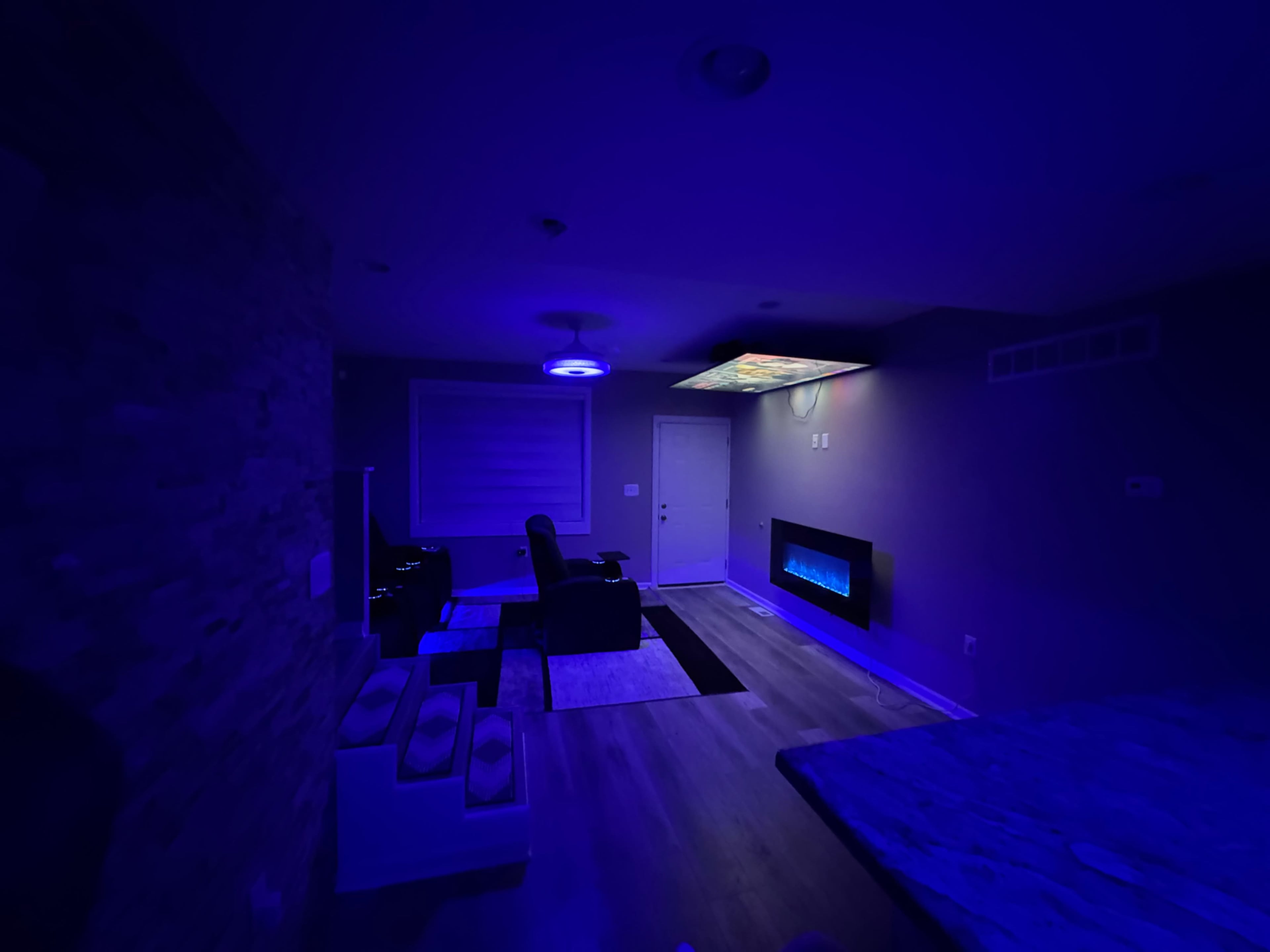 The image shows a dimly lit room with blue lighting, featuring a modern seating area and a wall-mounted electric fireplace.