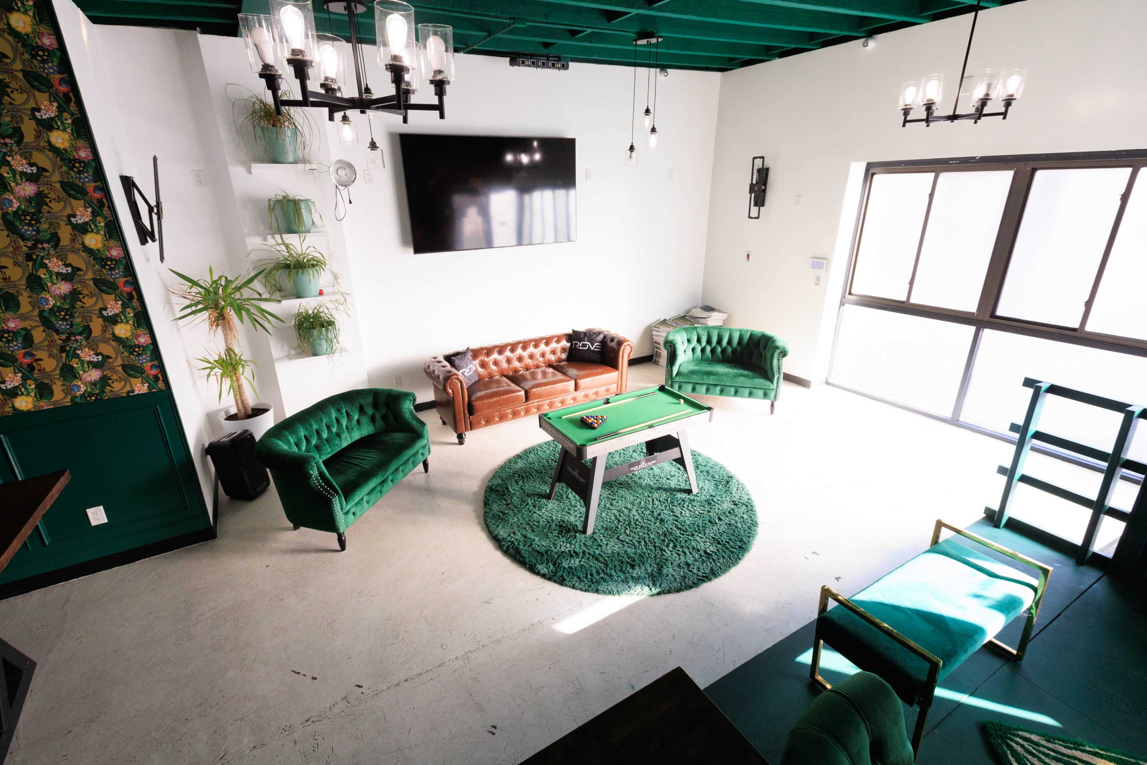 The image shows a modern living space featuring a brown leather sofa, green velvet chairs, a green pool table, and large windows allowing natural light.