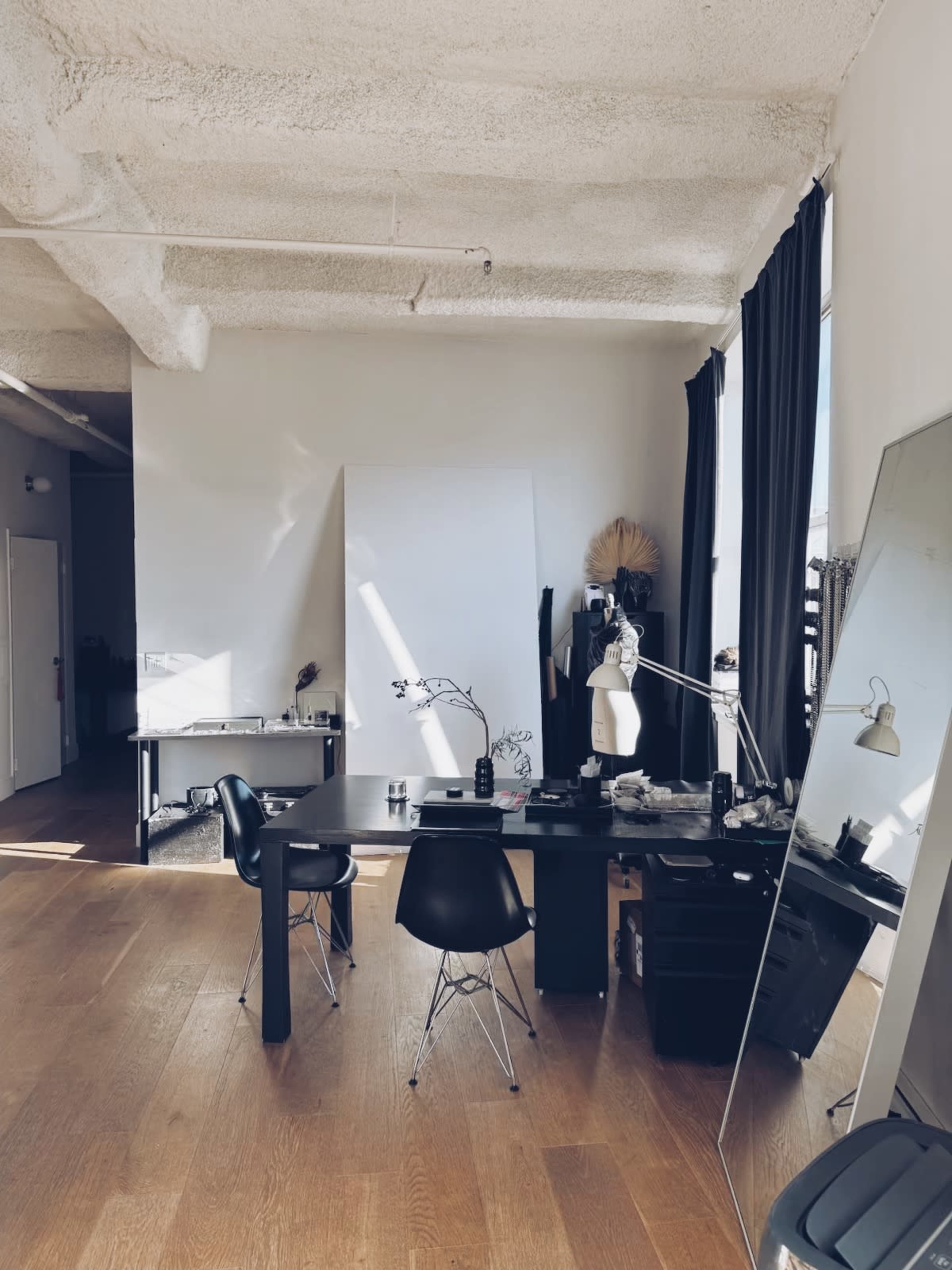 The image shows a minimalist studio space with a large window, a wooden floor, a desk with a lamp and chairs, and a wall featuring a drawing.