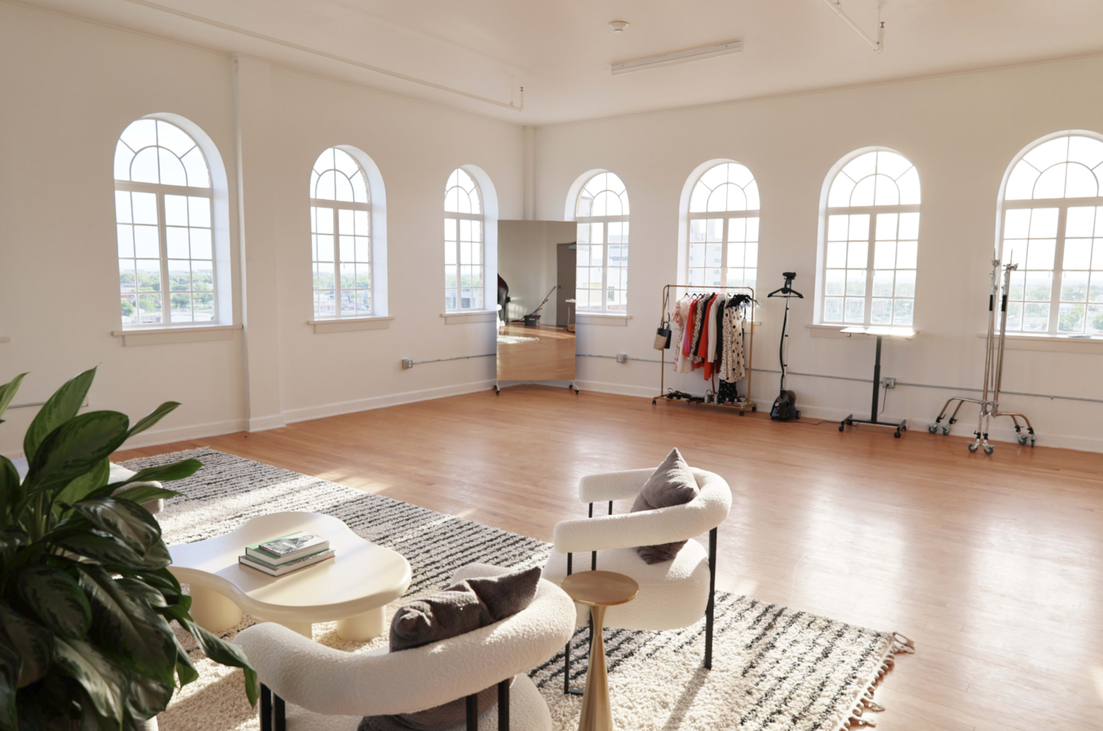A spacious room with tall arched windows, wooden flooring, and two chairs facing a small table, with clothing racks and a mirror in the background.