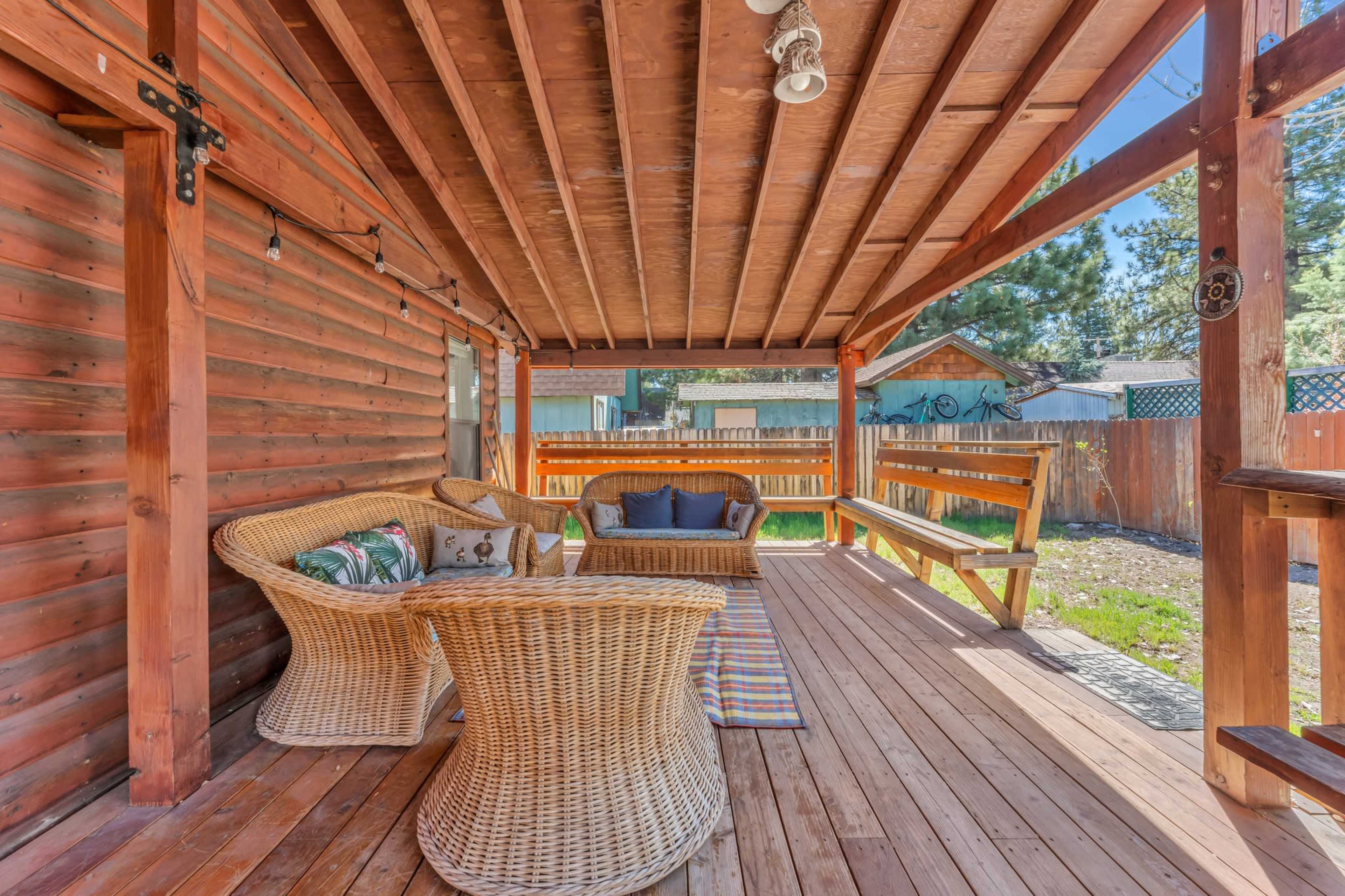 The image shows a spacious wooden porch featuring wicker furniture, a decorative rug, and a view of a grassy yard enclosed by a wooden fence.