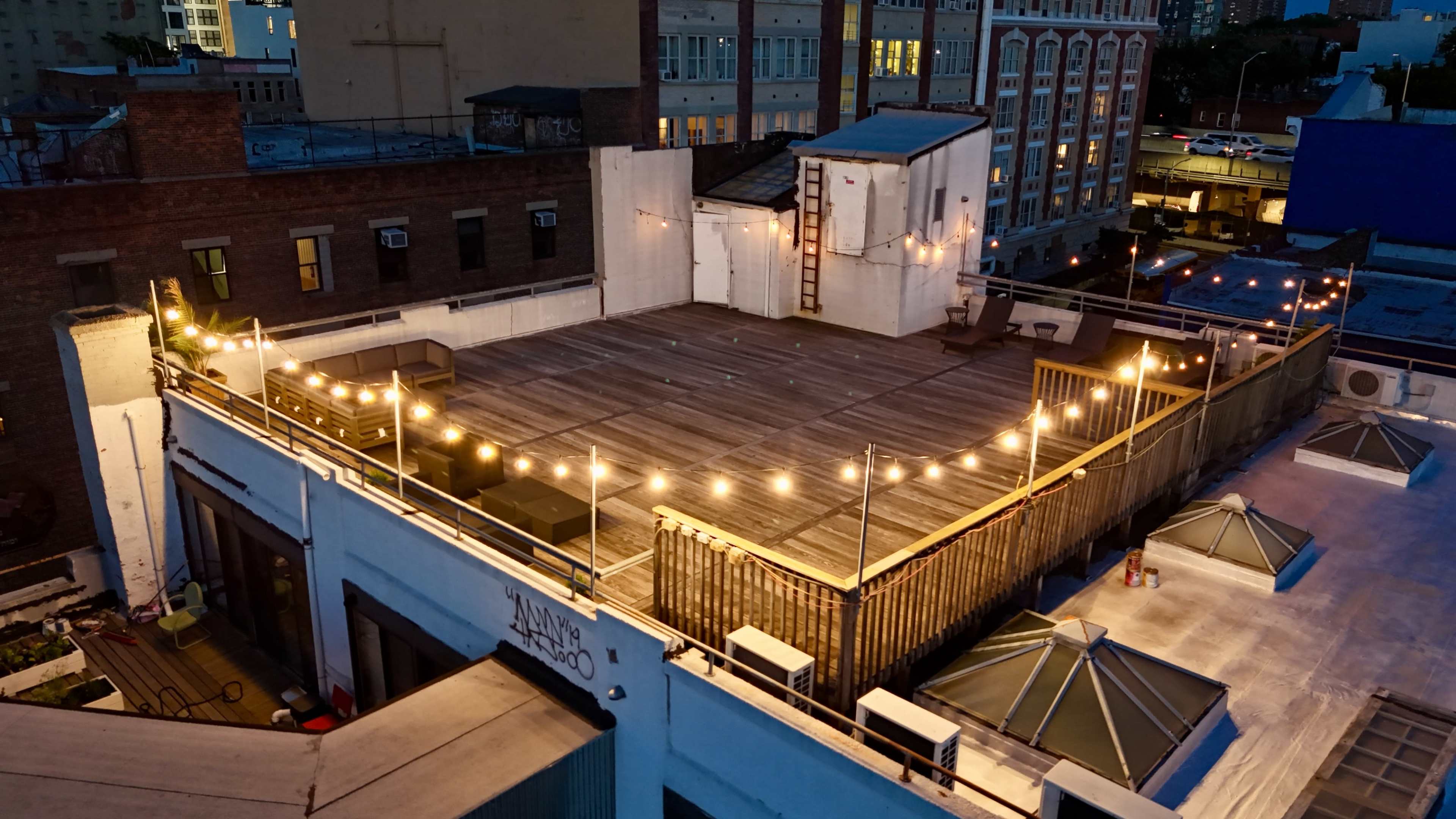 A rooftop terrace with wooden flooring illuminated by string lights, surrounded by buildings in an urban setting.
