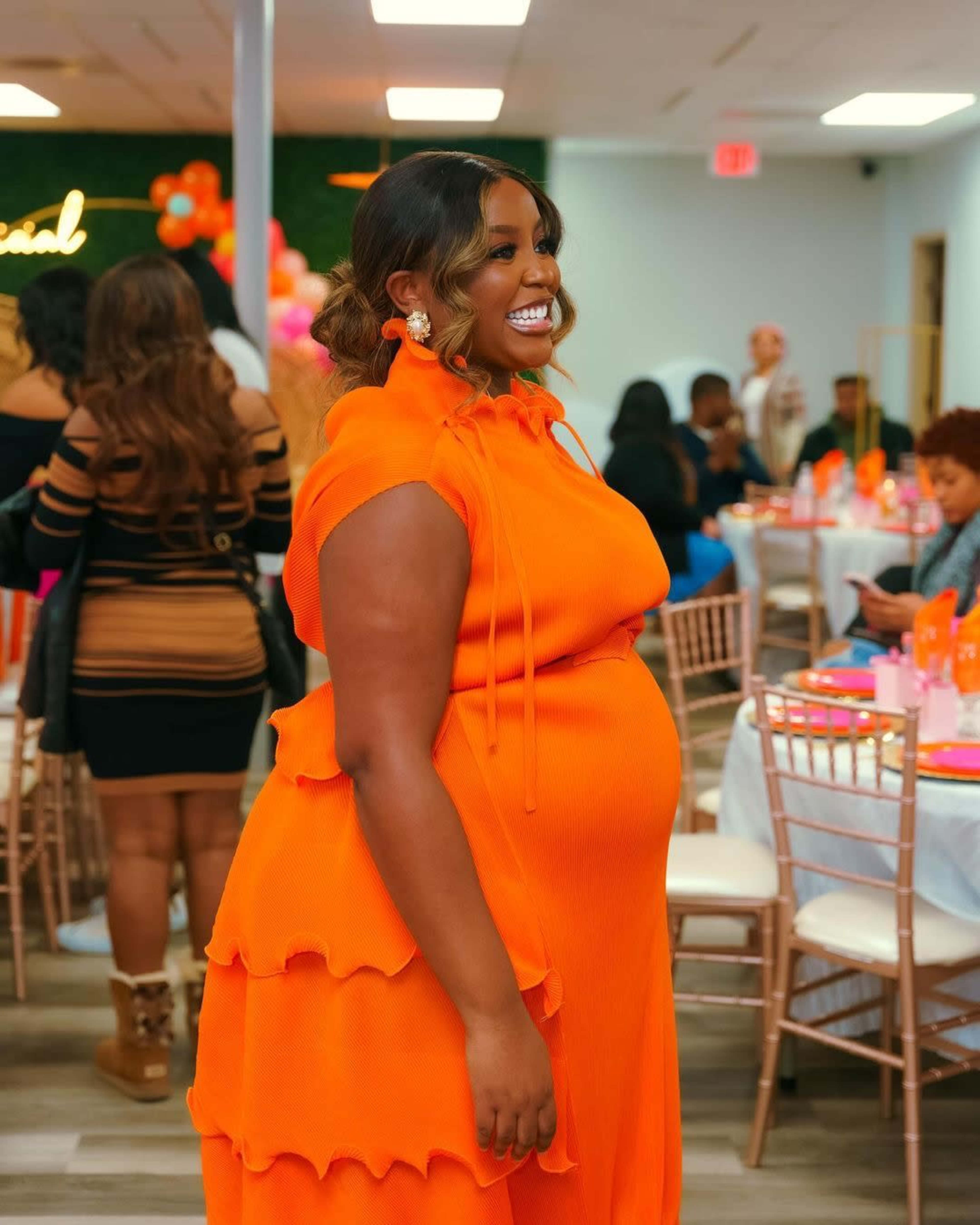 A woman in an orange dress stands smiling in a decorated event space filled with colorful table settings and guests.