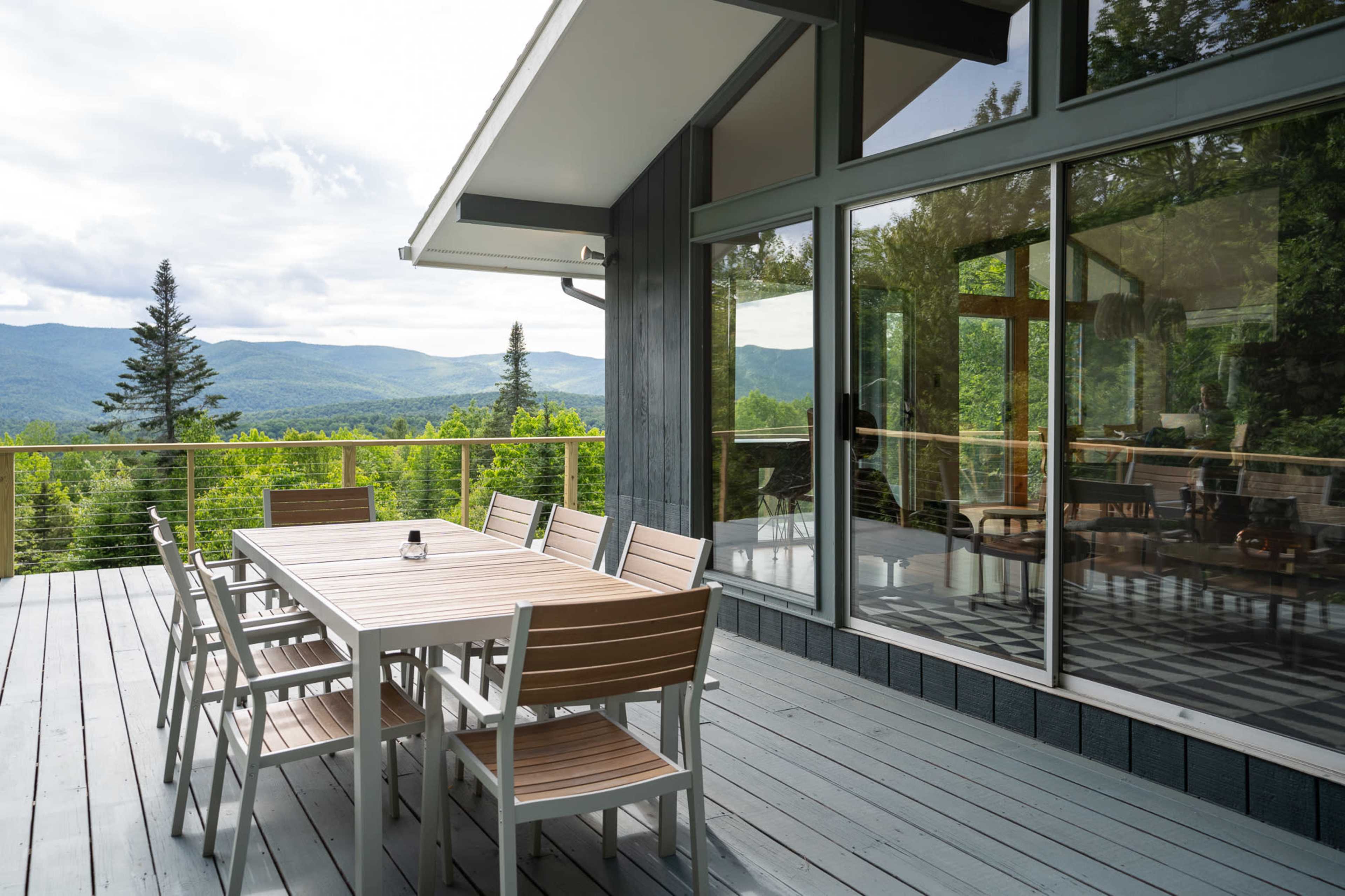 A modern wooden deck features a large dining table surrounded by chairs, with a view of mountains in the background and large glass windows reflecting the scenery.