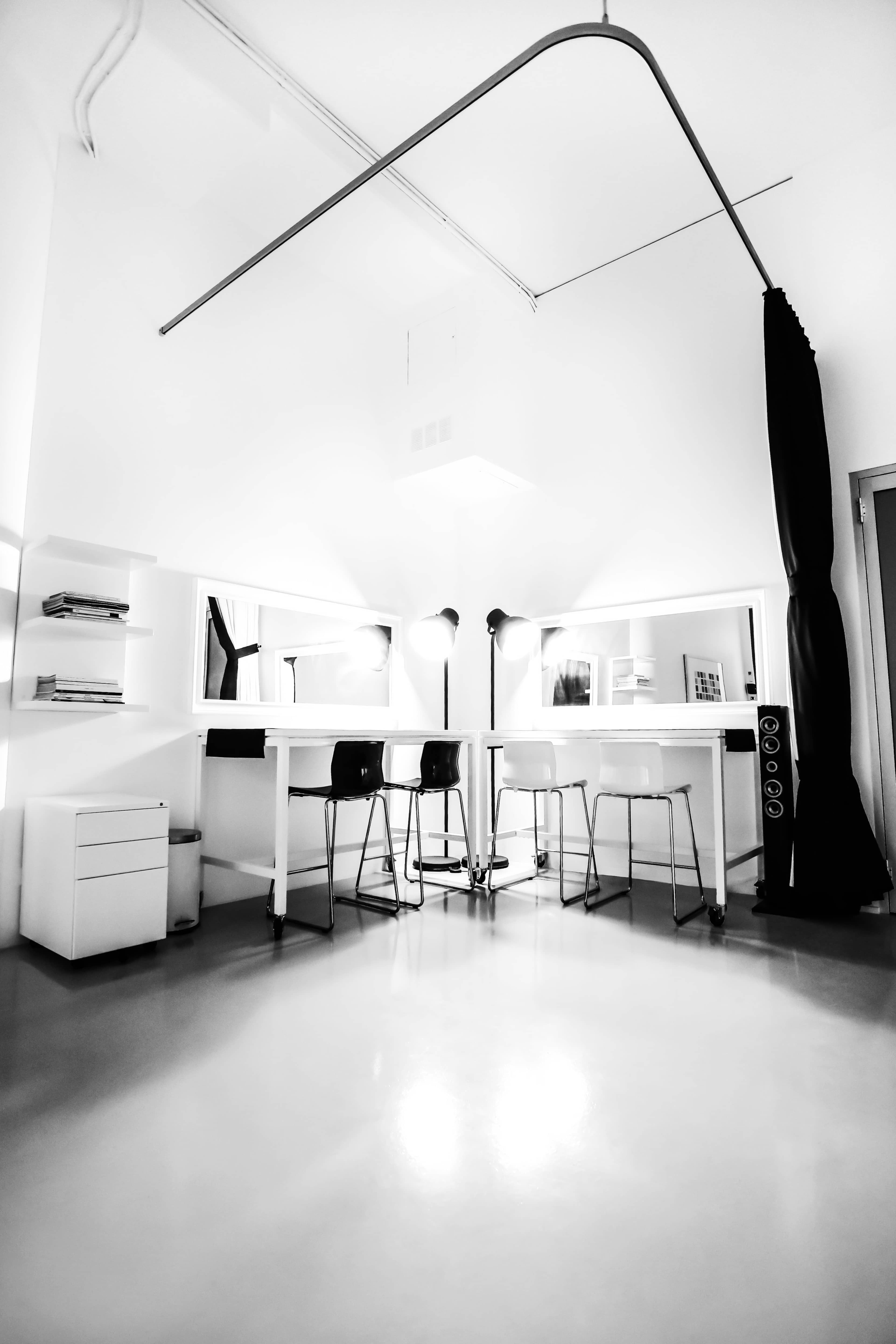 A minimalistic workspace with a white table and several high stools, illuminated by overhead lights in a room with white walls and a dark curtain.