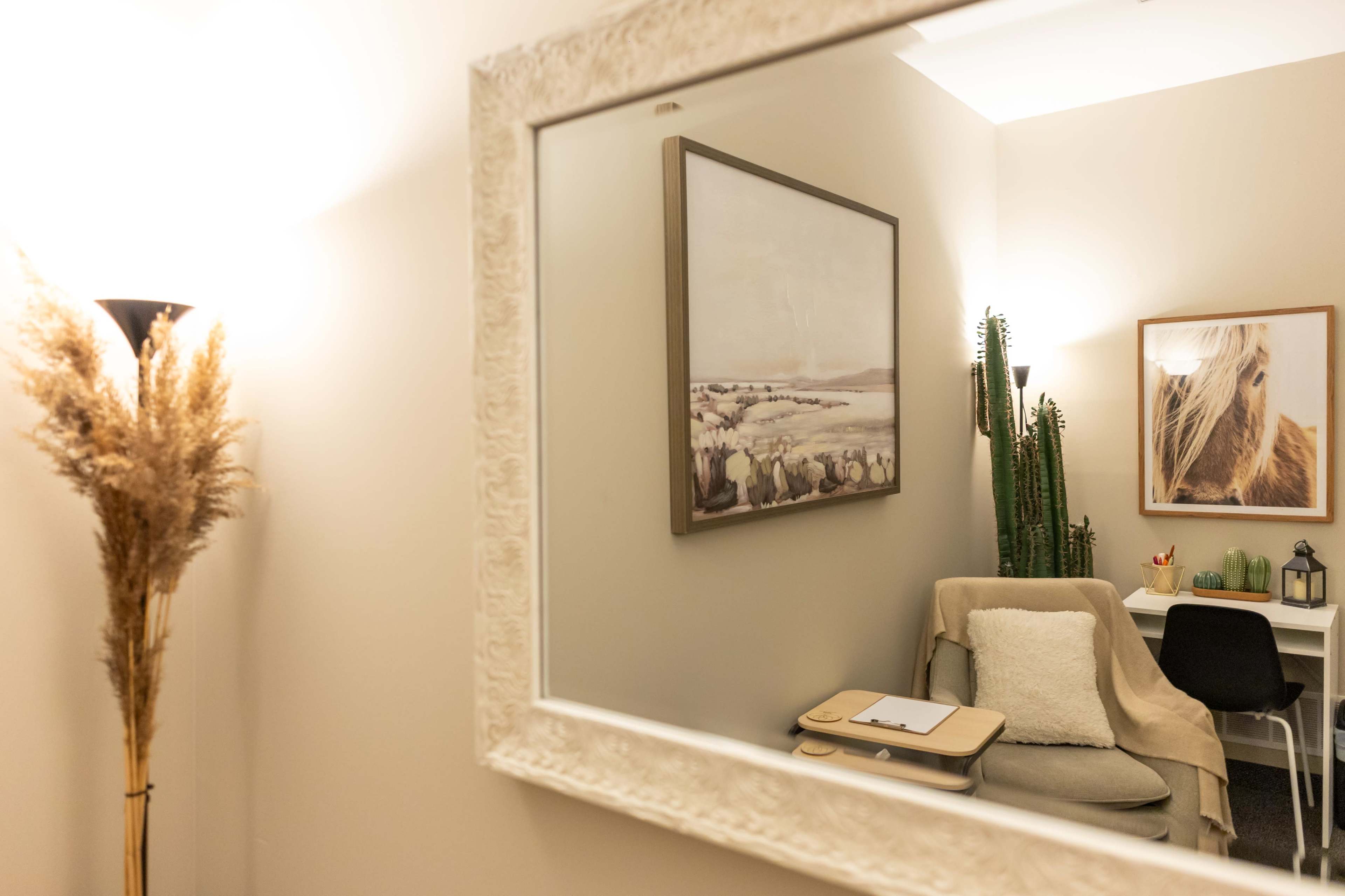 The image shows a cozy room featuring a mirror reflecting a framed painting, a desk with decorative items, a chair with a blanket, and a tall plant in the corner.