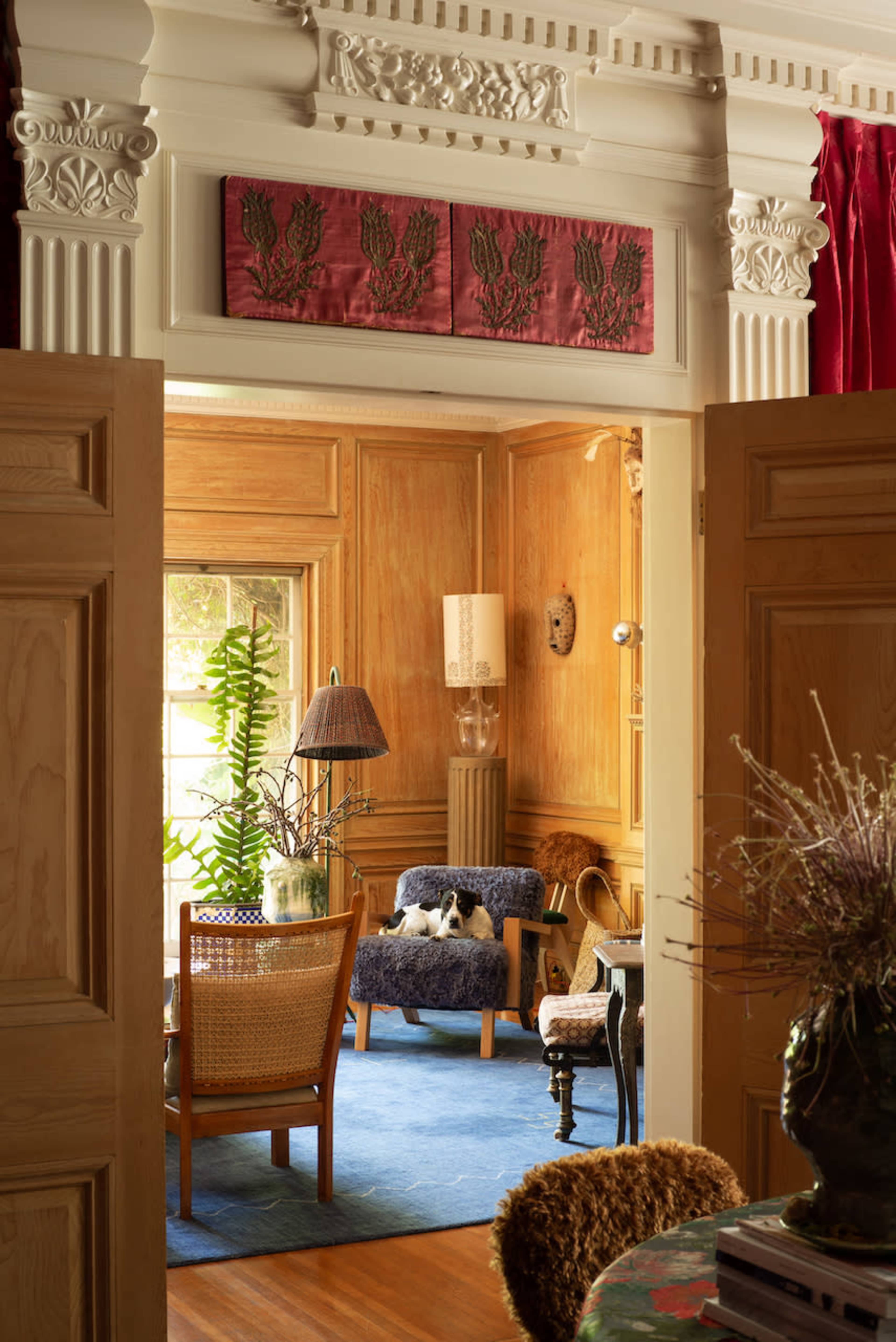 The image depicts a cozy interior space featuring wooden paneling, a decorative ceiling, and a seating area with a lamp and plants, viewed through an open door.