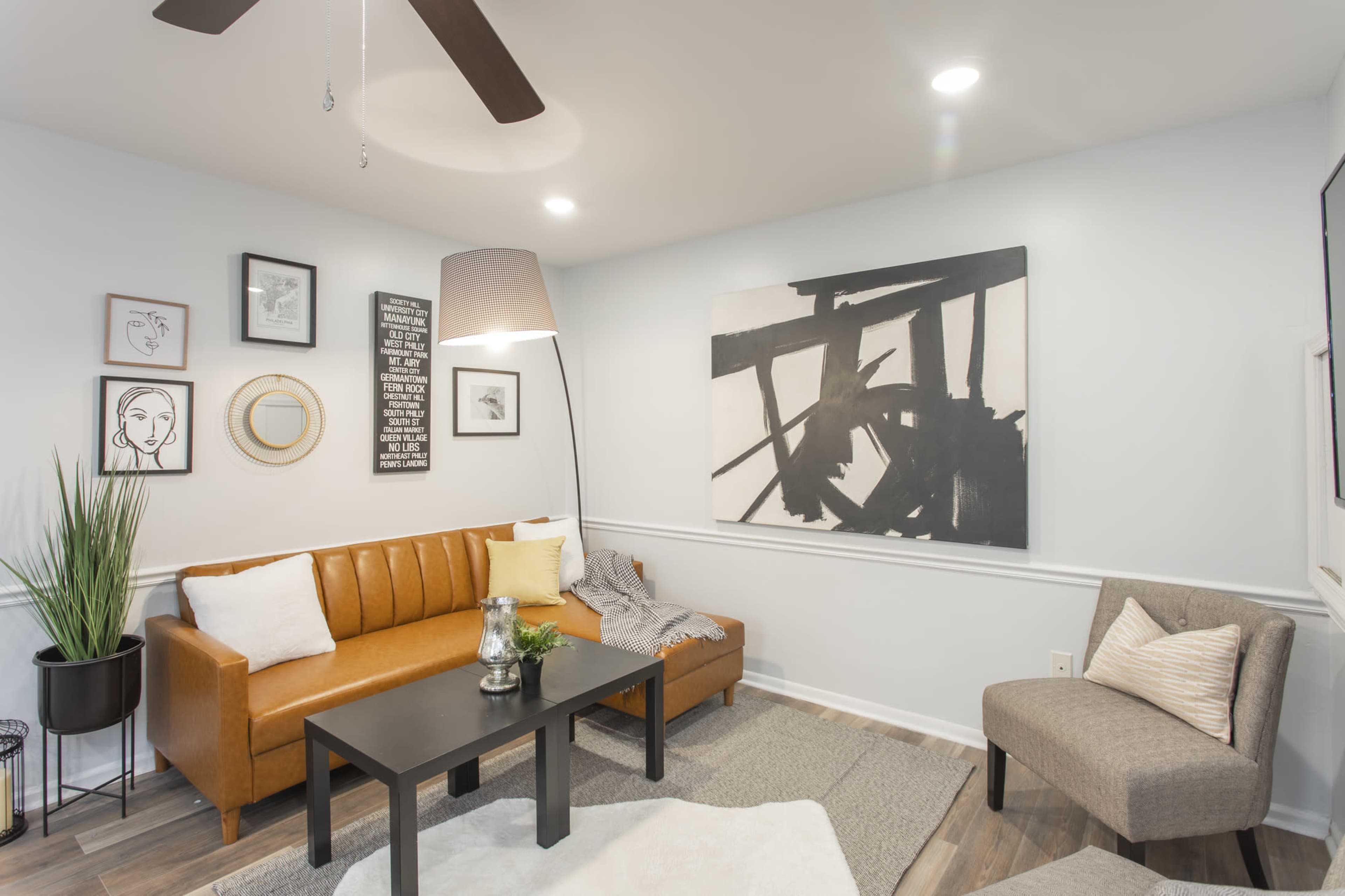 A modern living room features a brown leather sofa, a black coffee table, artwork on the walls, and a gray chair, all arranged with a light rug on a wooden floor.