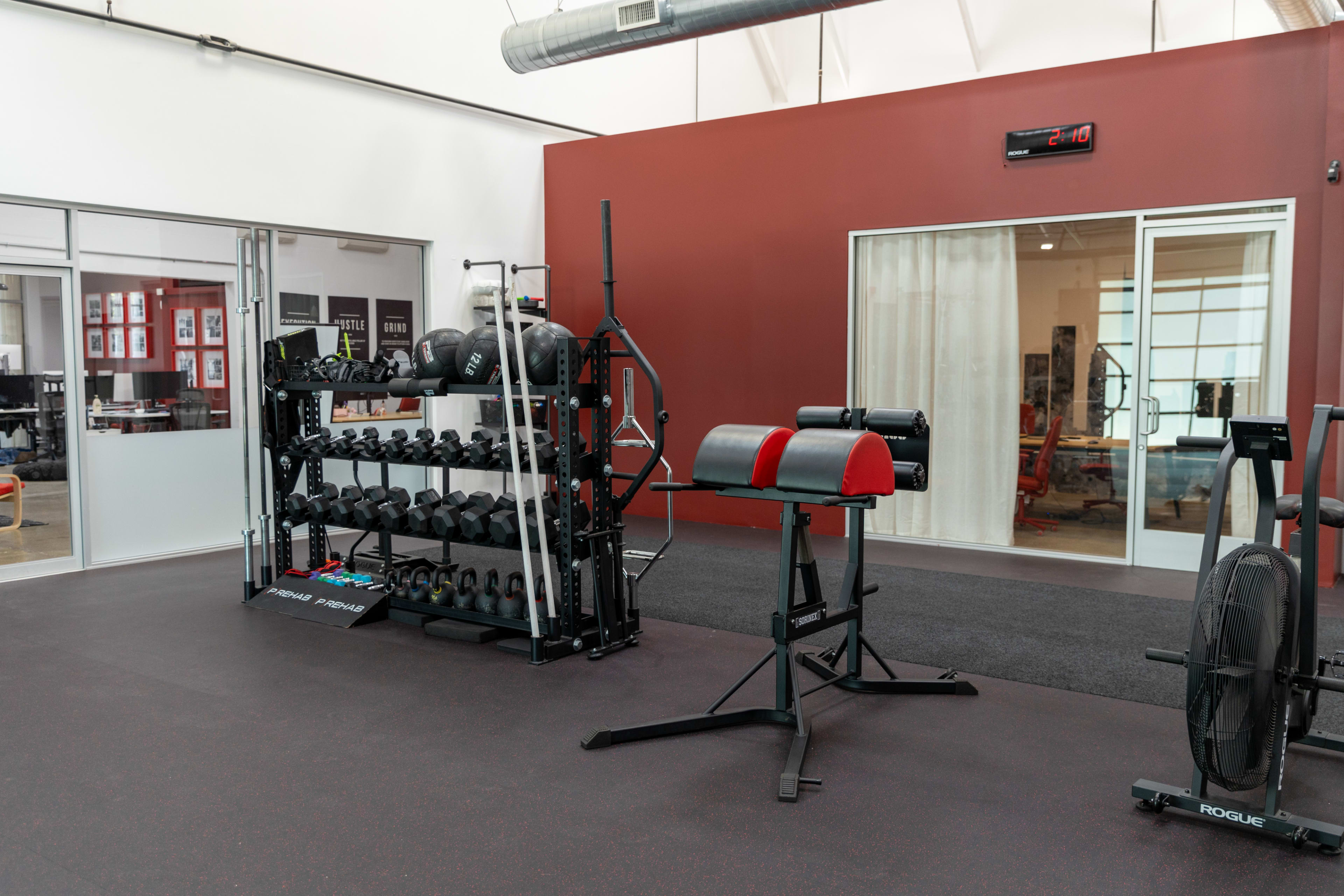 The image shows a well-equipped gym area featuring a rack of dumbbells, a weight bench, and a stationary bike on a rubber floor.