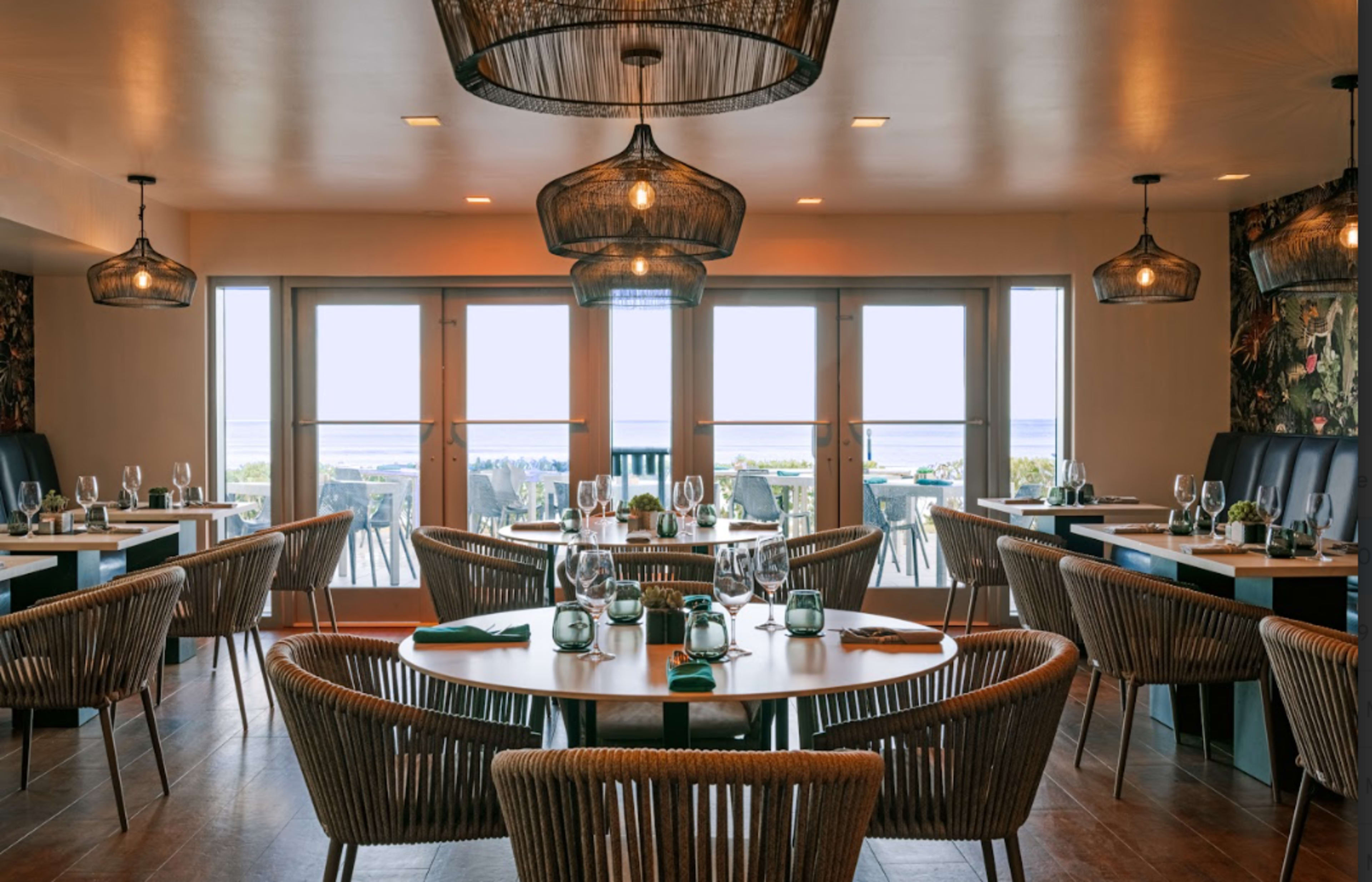 The image shows a modern restaurant interior with round tables, wicker chairs, and large windows offering a view of the outdoors.