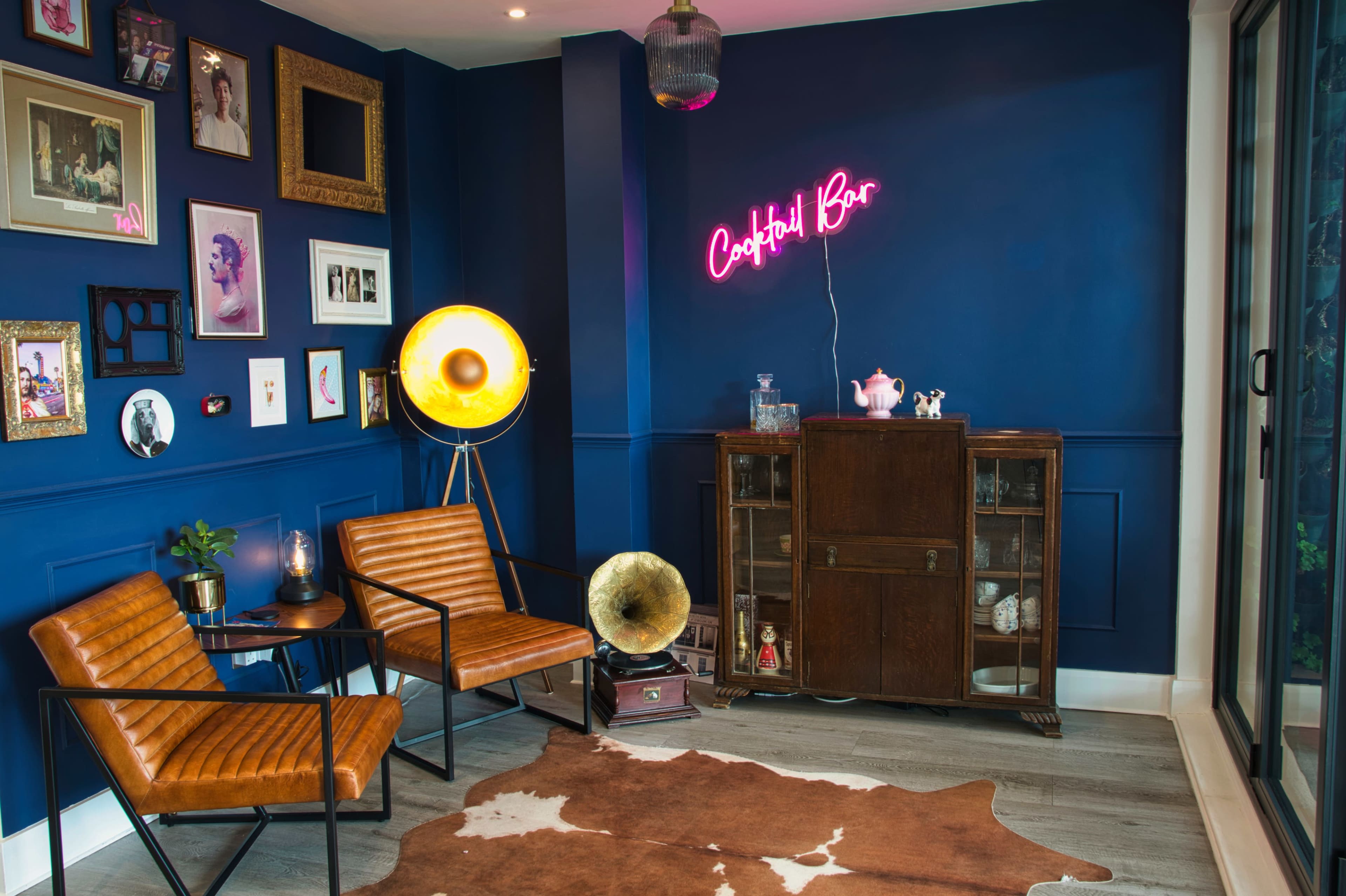 A cozy lounge area features leather chairs, a vintage gramophone, a neon sign reading "Cocktail Bar," and framed artwork on a deep blue wall.