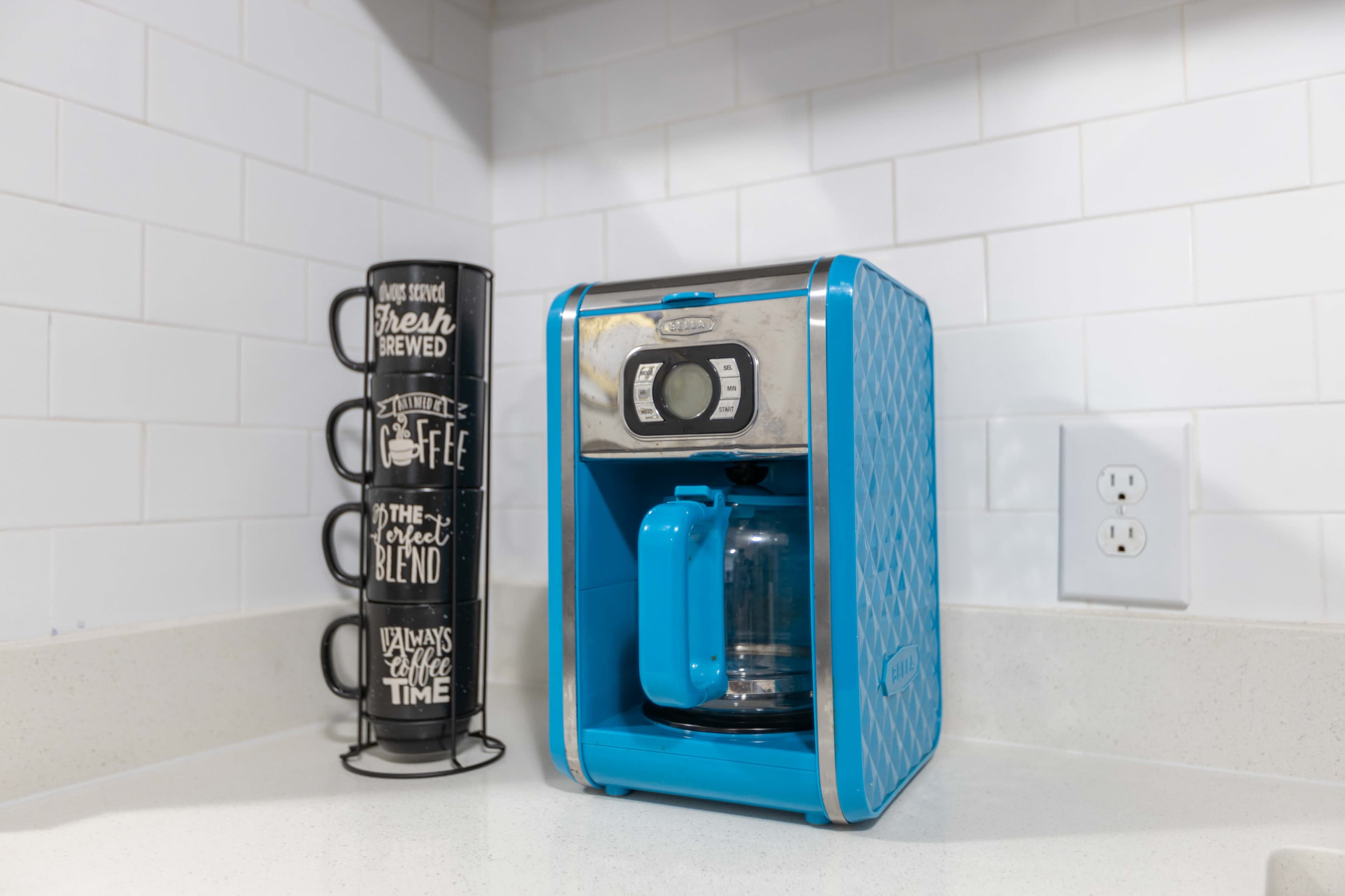 A blue coffee maker sits on a countertop next to a stack of black coffee mugs.