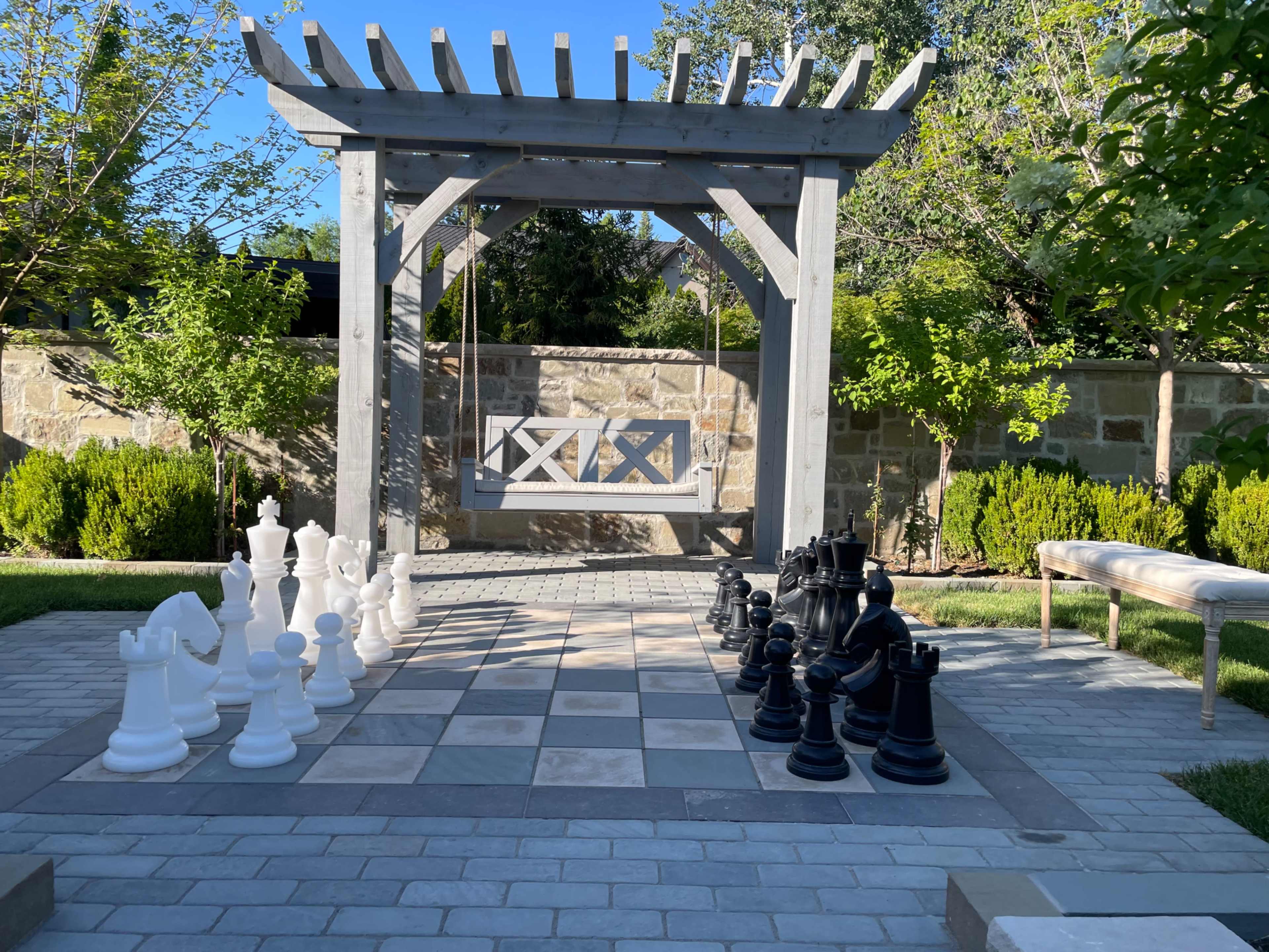 A large outdoor chessboard with black and white pieces is set in a garden featuring a wooden swing beneath a pergola.
