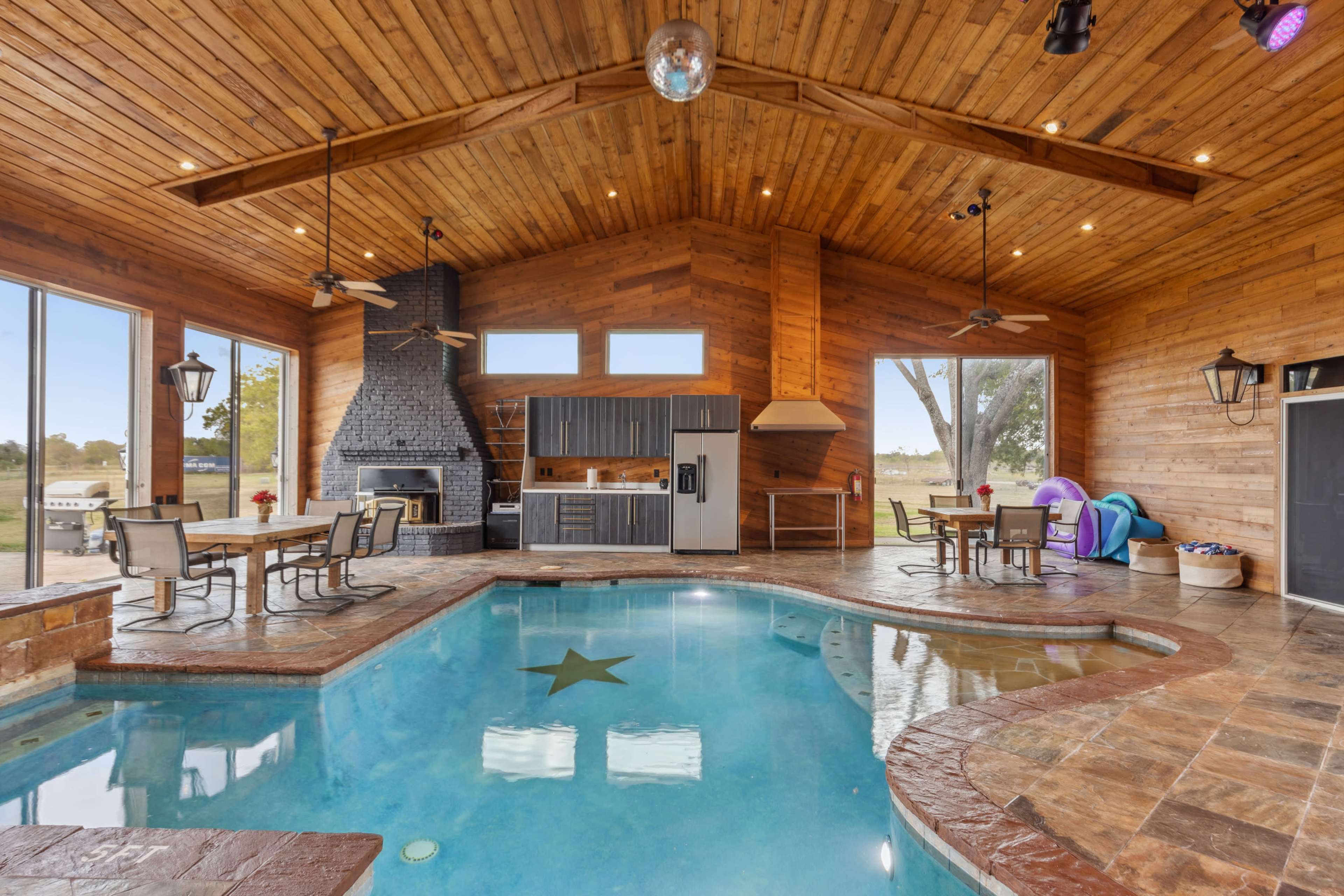 The image shows a wooden-paneled indoor space featuring a swimming pool, a brick fireplace, and a kitchenette, with large windows overlooking a grassy area.