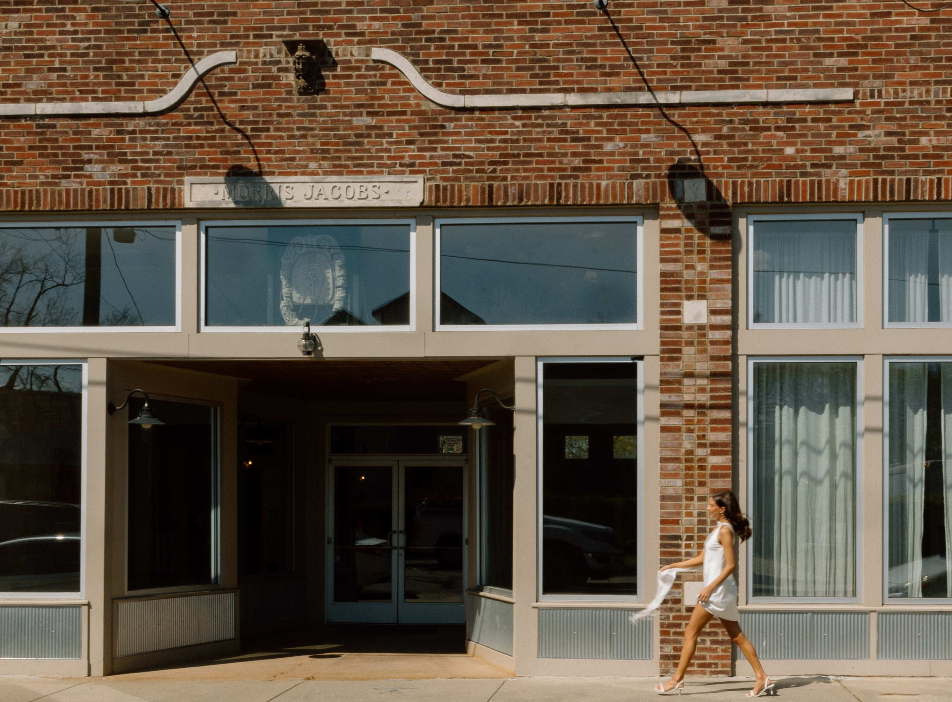 A woman in a white dress walks past a brick building with large windows and an entrance labeled "Adams Jacobs."