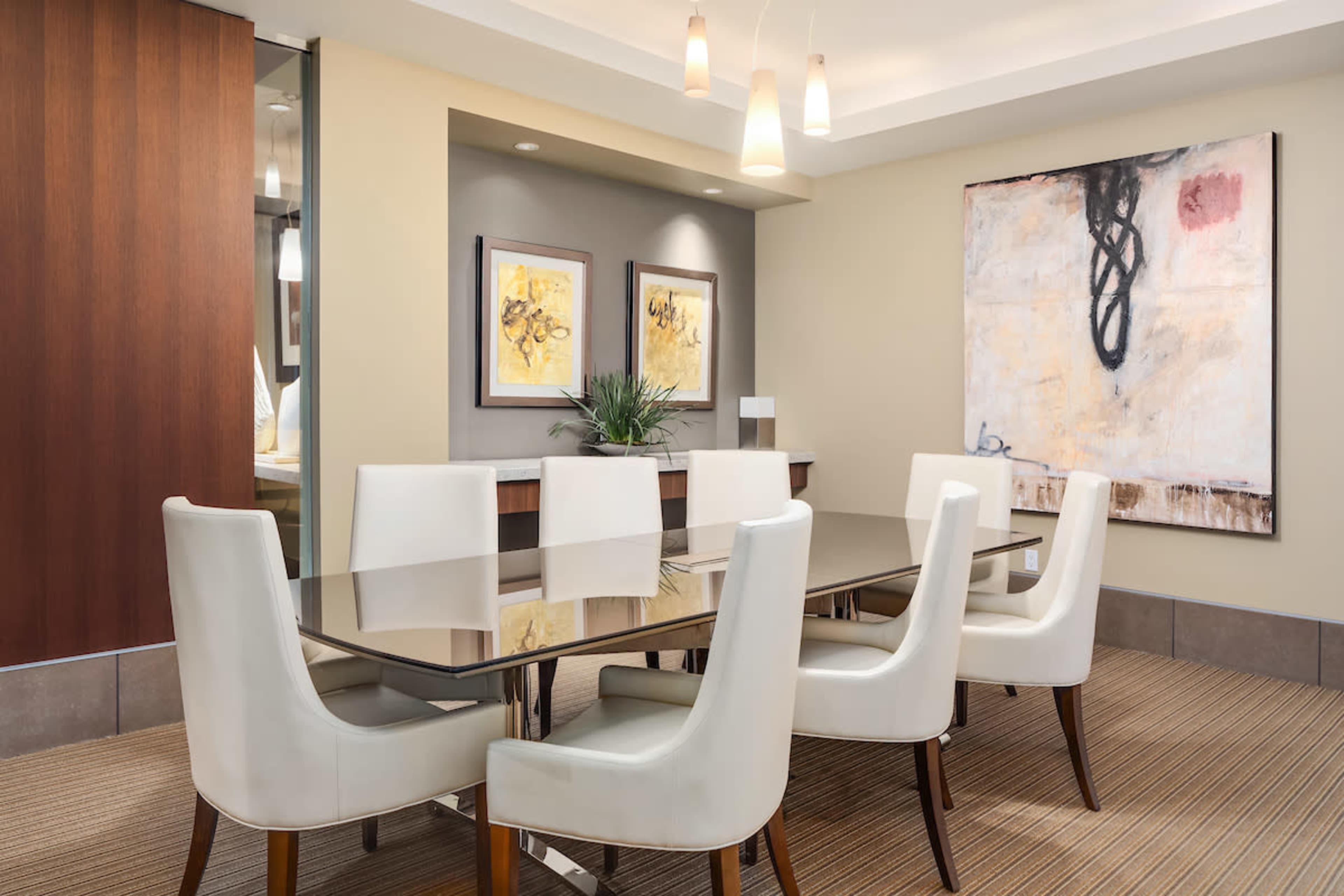 A modern conference room features a long glass table surrounded by eight white chairs, with artwork on the walls and a plant on a side table.