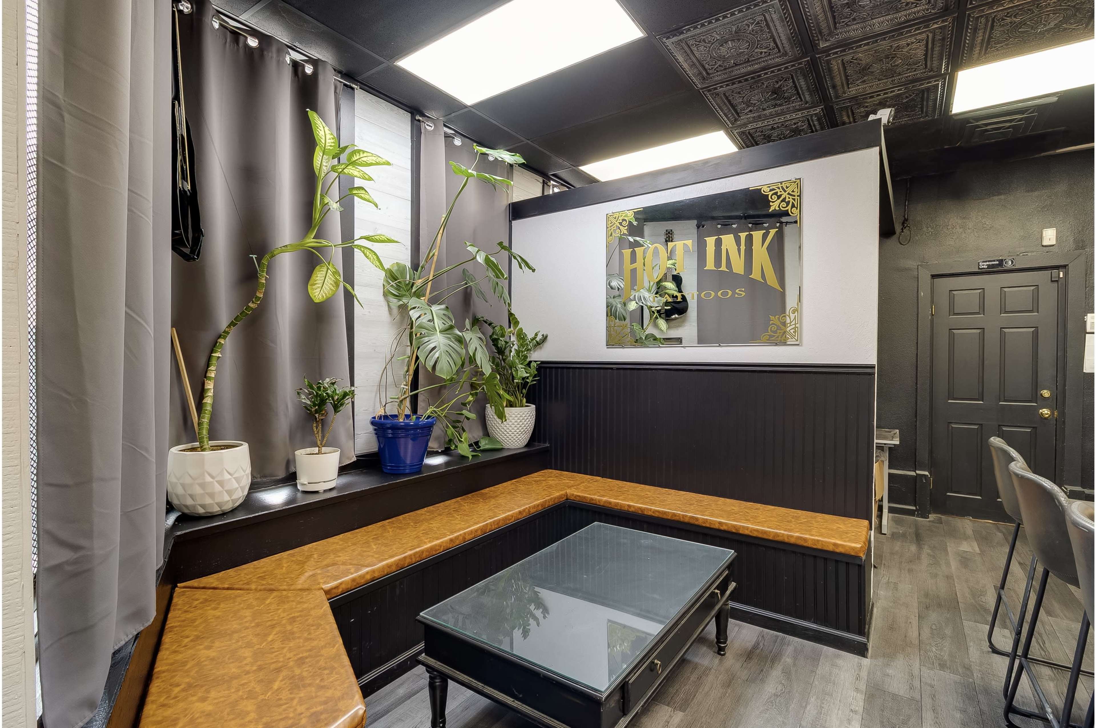 The image shows a modern waiting area in a tattoo studio, featuring a seating area with a wooden bench, a glass coffee table, and several potted plants.