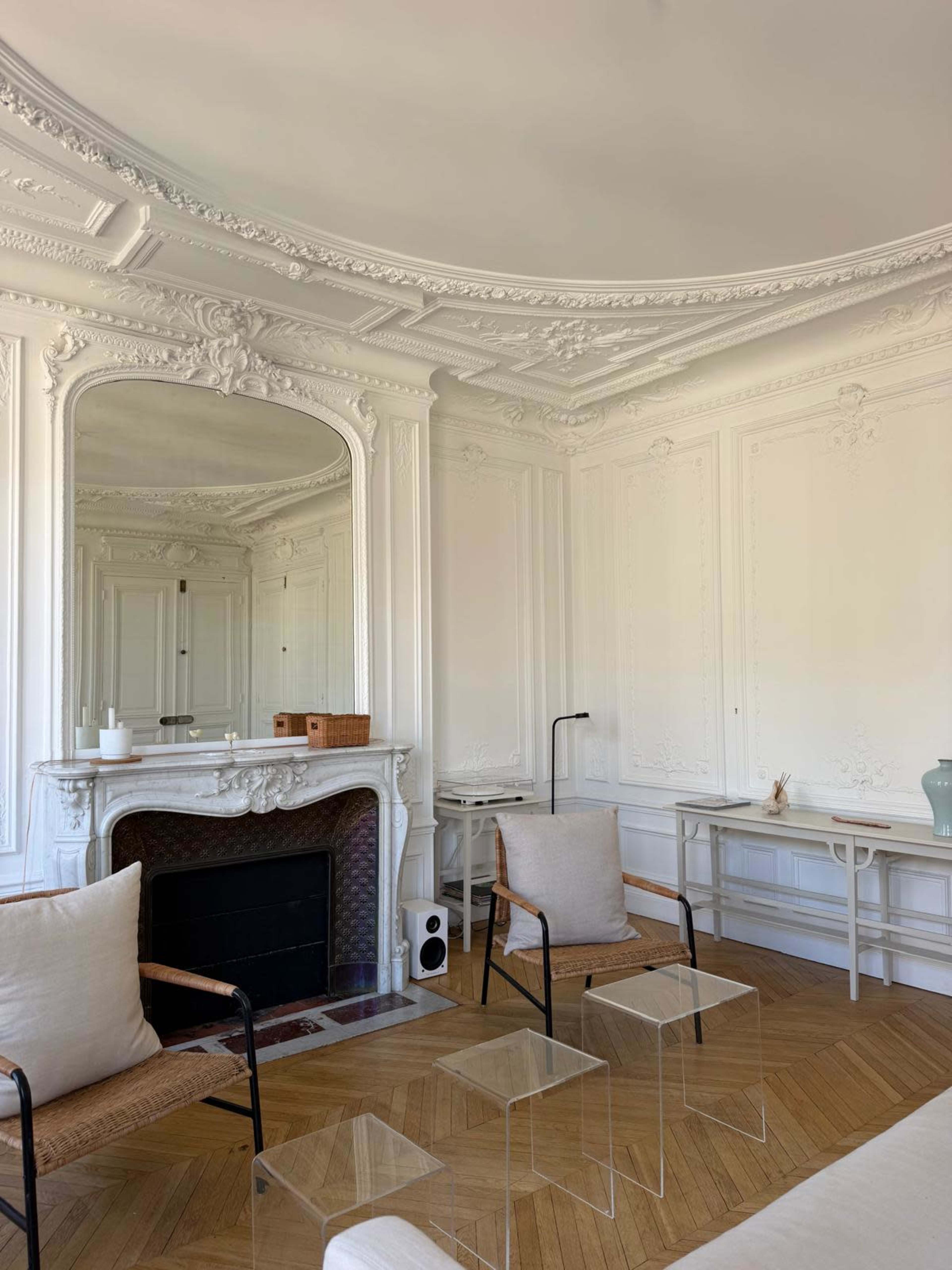 The image shows a bright room with intricate white moldings, a large mirror above a decorative fireplace, and transparent chairs arranged around a minimalist table.