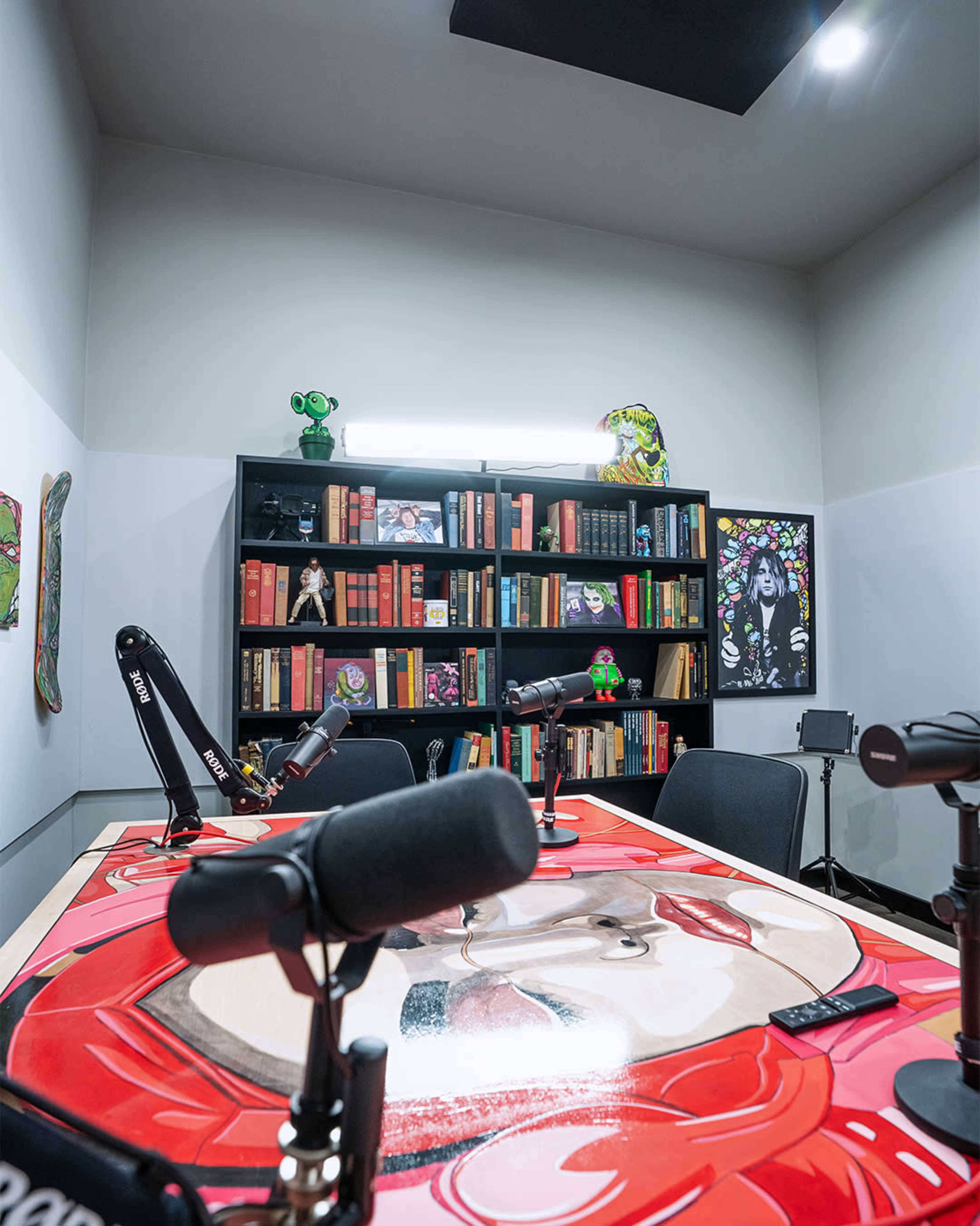 A podcast recording studio with a circular table, multiple microphones, and a bookshelf filled with colorful books and decorative items on the walls.