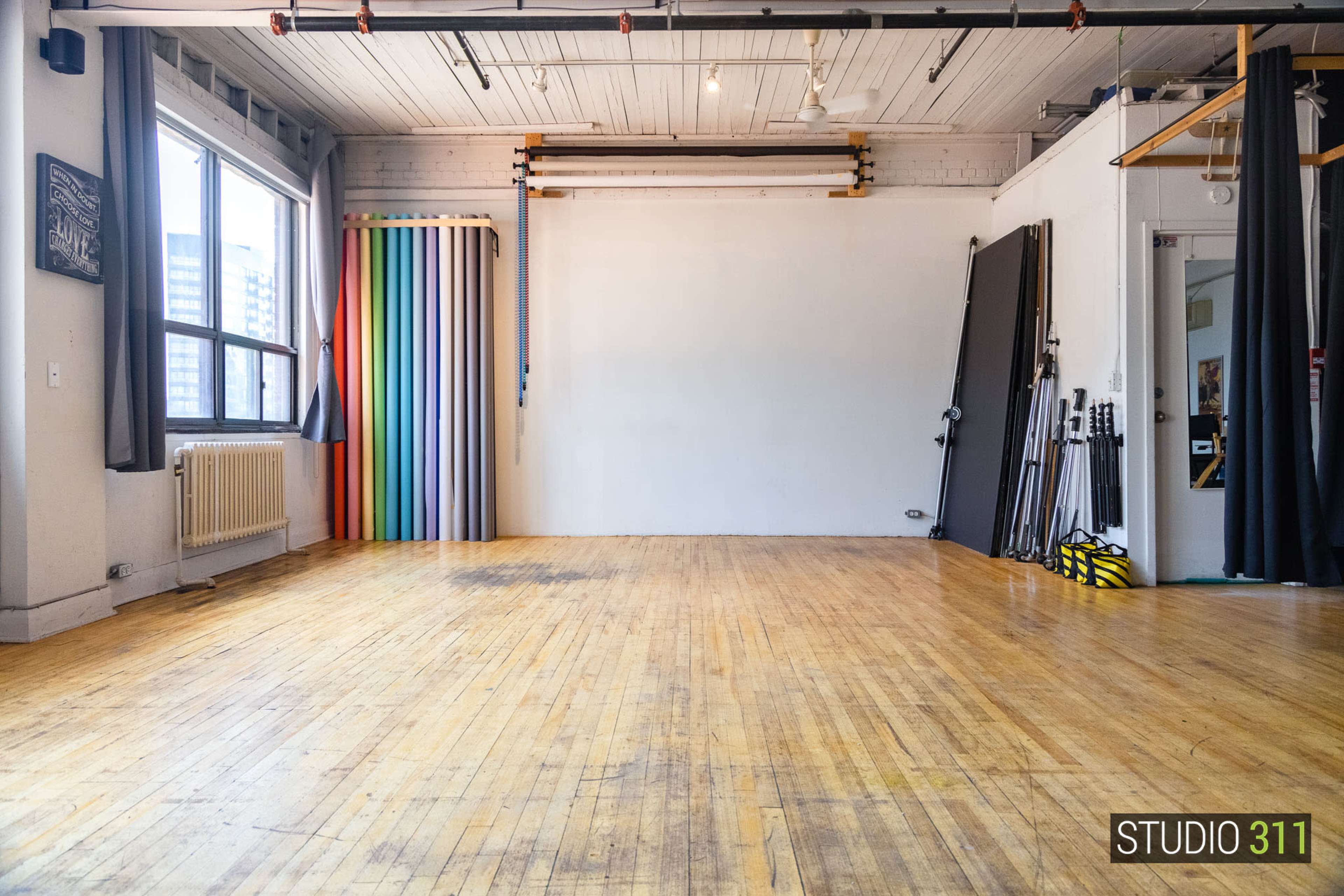 The image shows a spacious studio with wooden floors, large windows, and various colorful backdrops stacked against the wall.