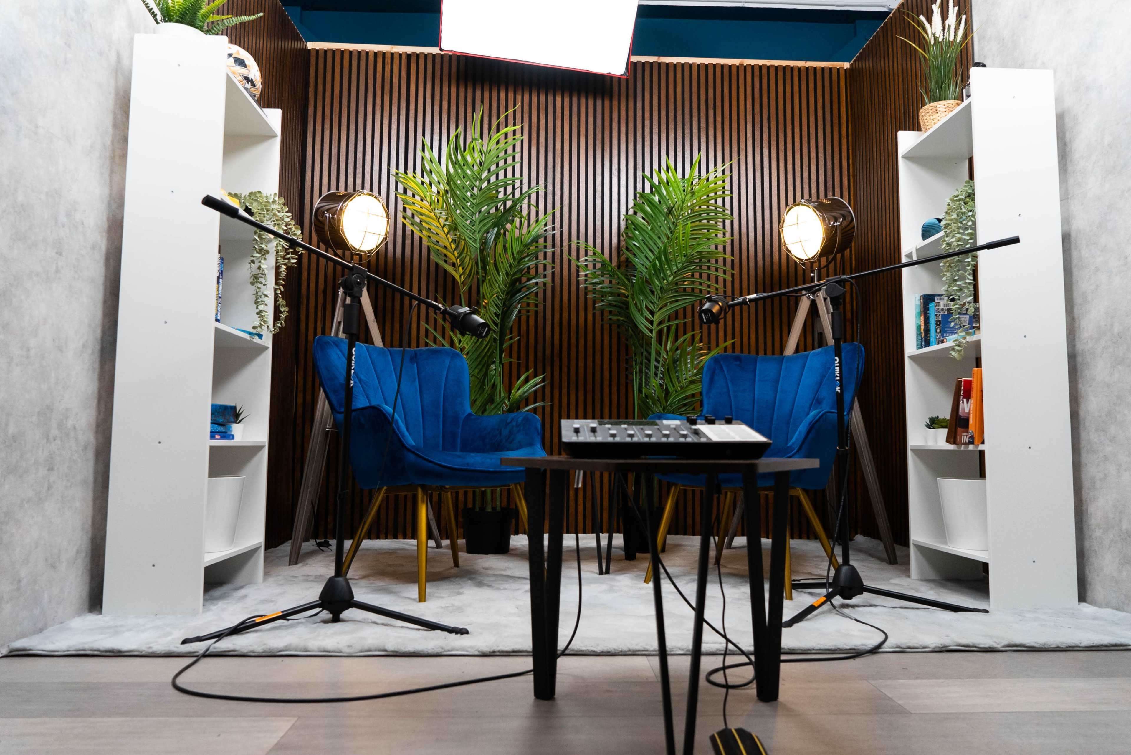 The scene features two blue chairs on a soft rug facing a small table, with recording equipment and decorative plants in a modern studio setup.