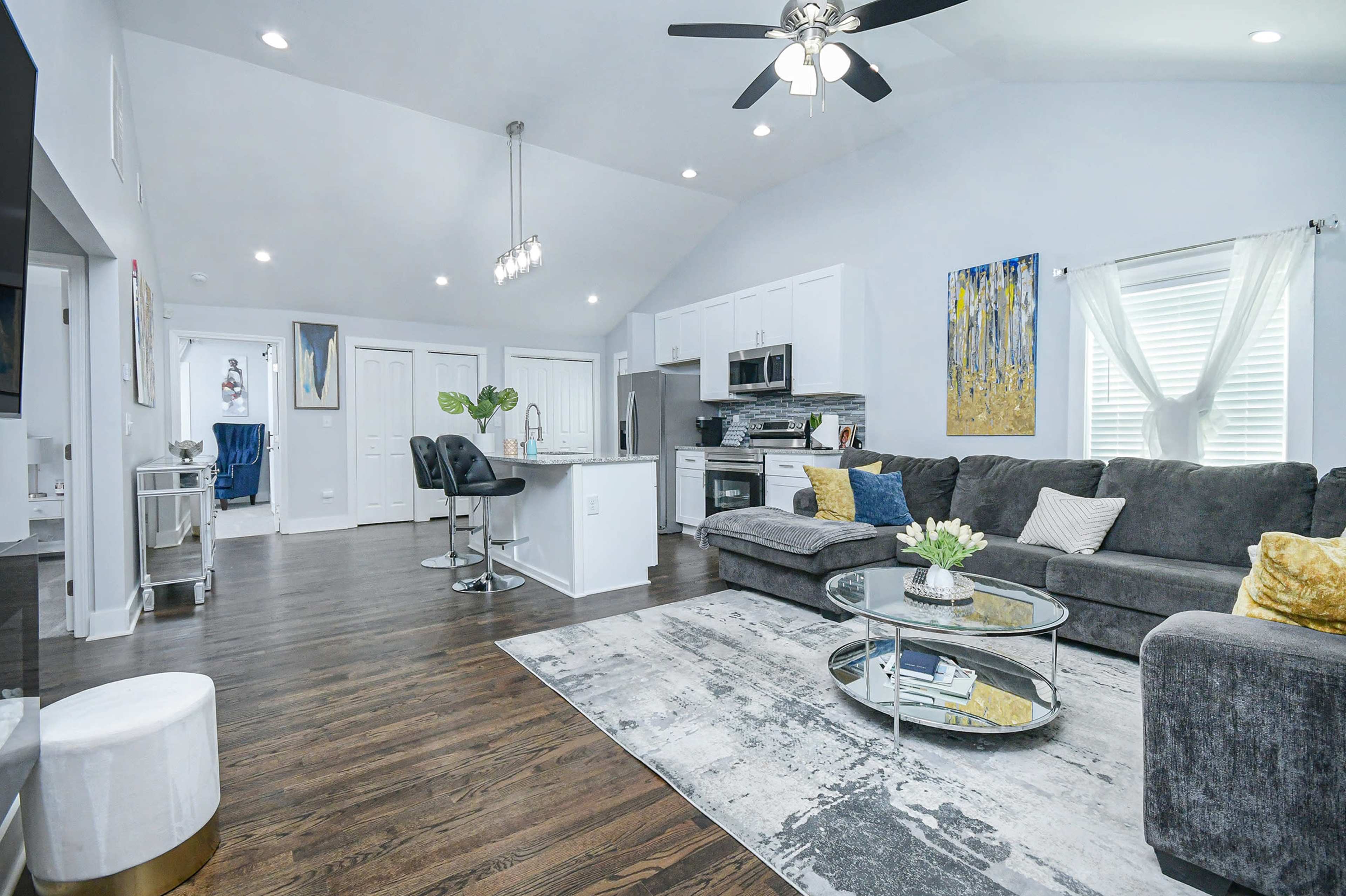 A modern living room with a sectional sofa, a round coffee table, a kitchen area, and large windows allowing natural light to enter.