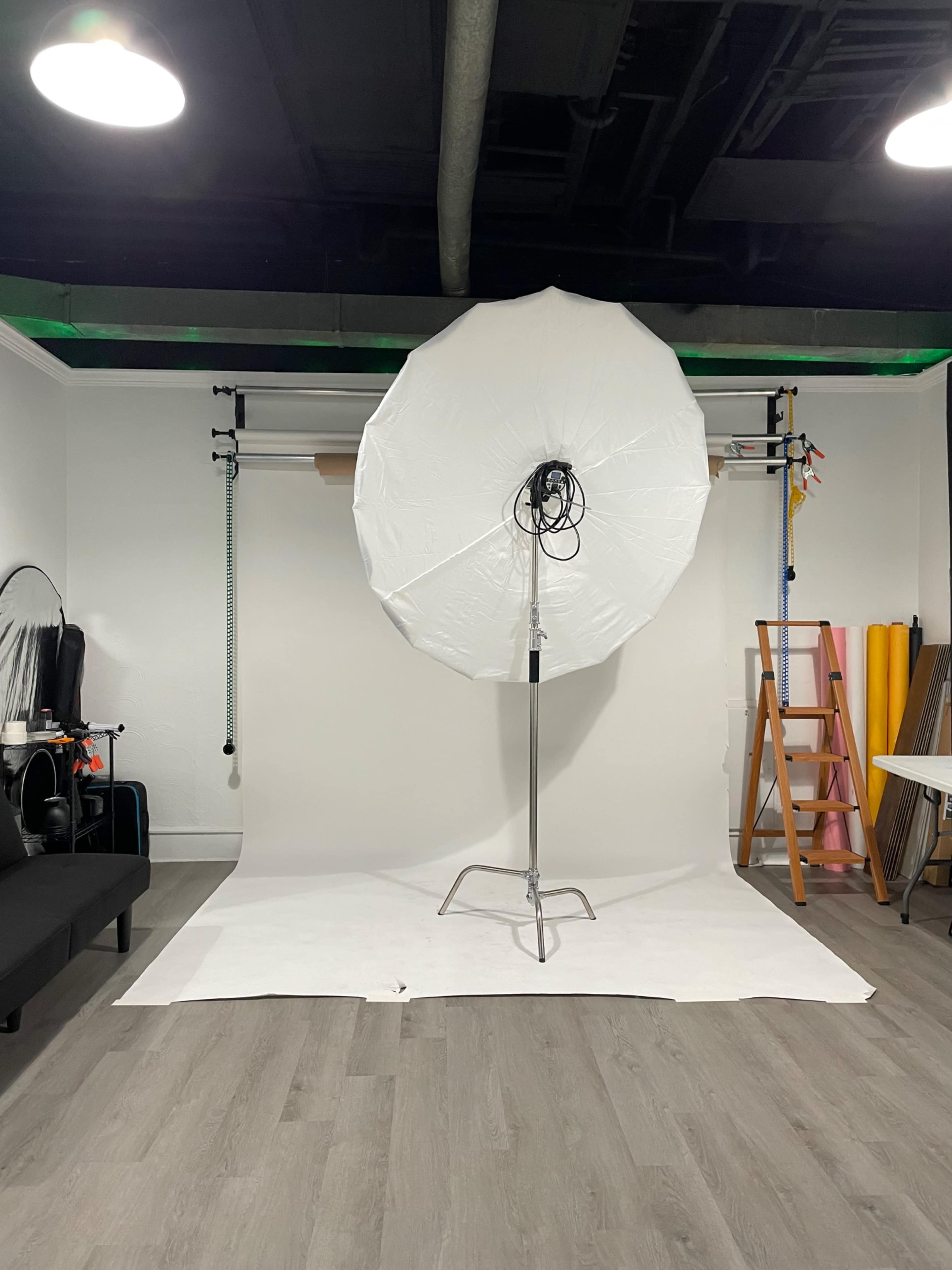 The image shows a photography studio with an umbrella softbox positioned in front of a plain backdrop, surrounded by various studio equipment and a ladder.