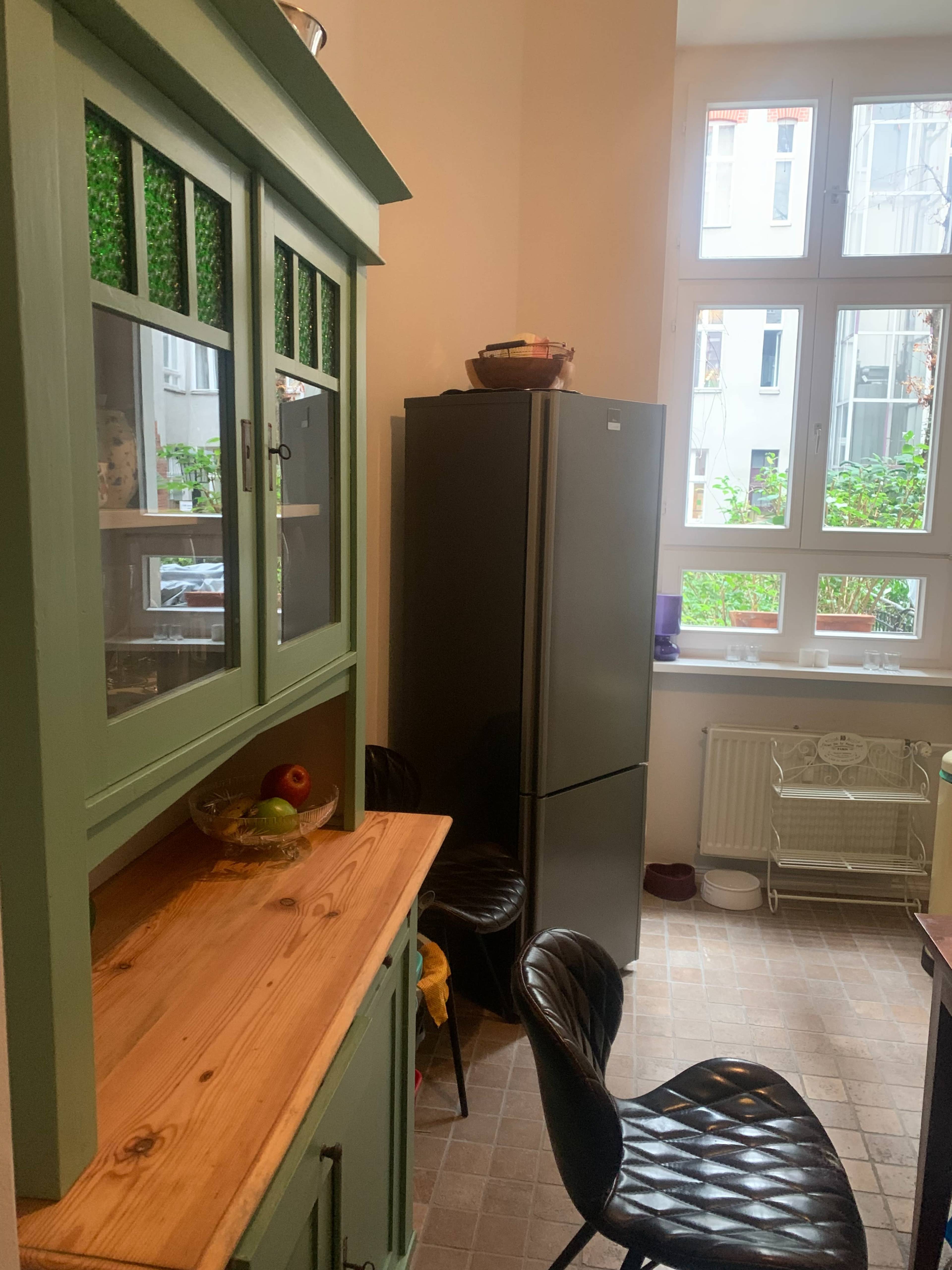 A modern kitchen features a green cabinet, a gray refrigerator, and black chairs arranged near a wooden countertop.