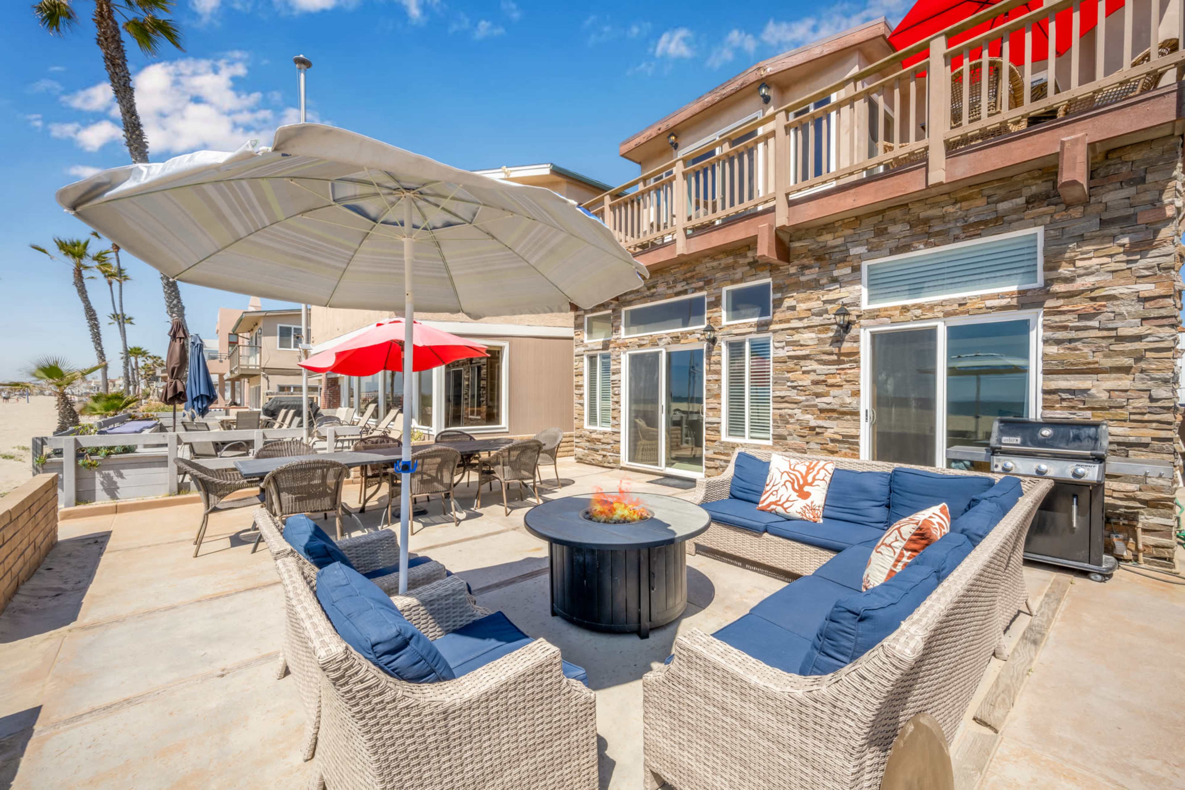 A patio area with wicker furniture, a round table, an outdoor grill, and large umbrellas, set in a coastal environment.