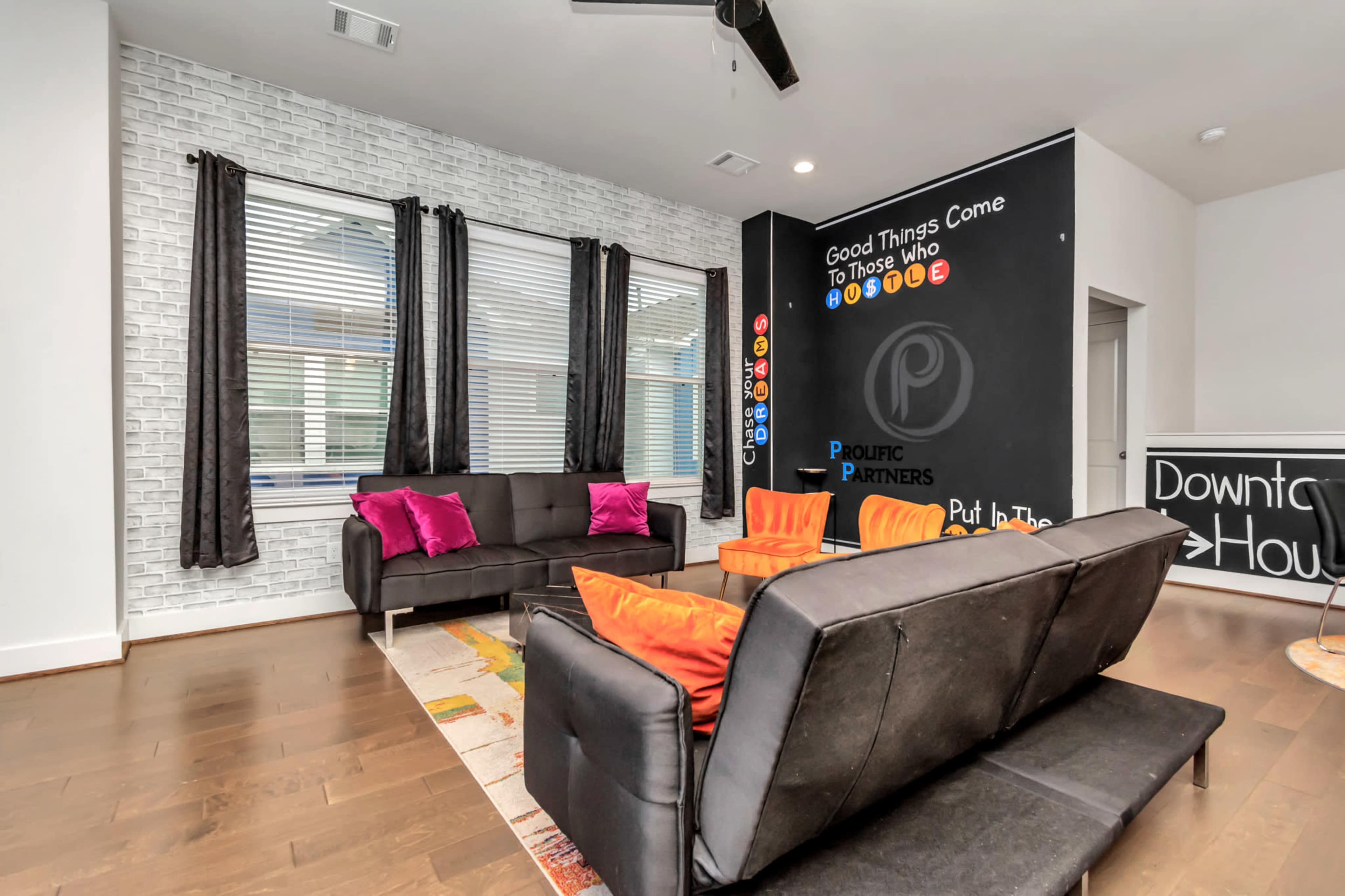 The image shows a modern living room with a black sofa, colorful chairs, a patterned rug, and a feature wall with motivational text and a chalkboard area.