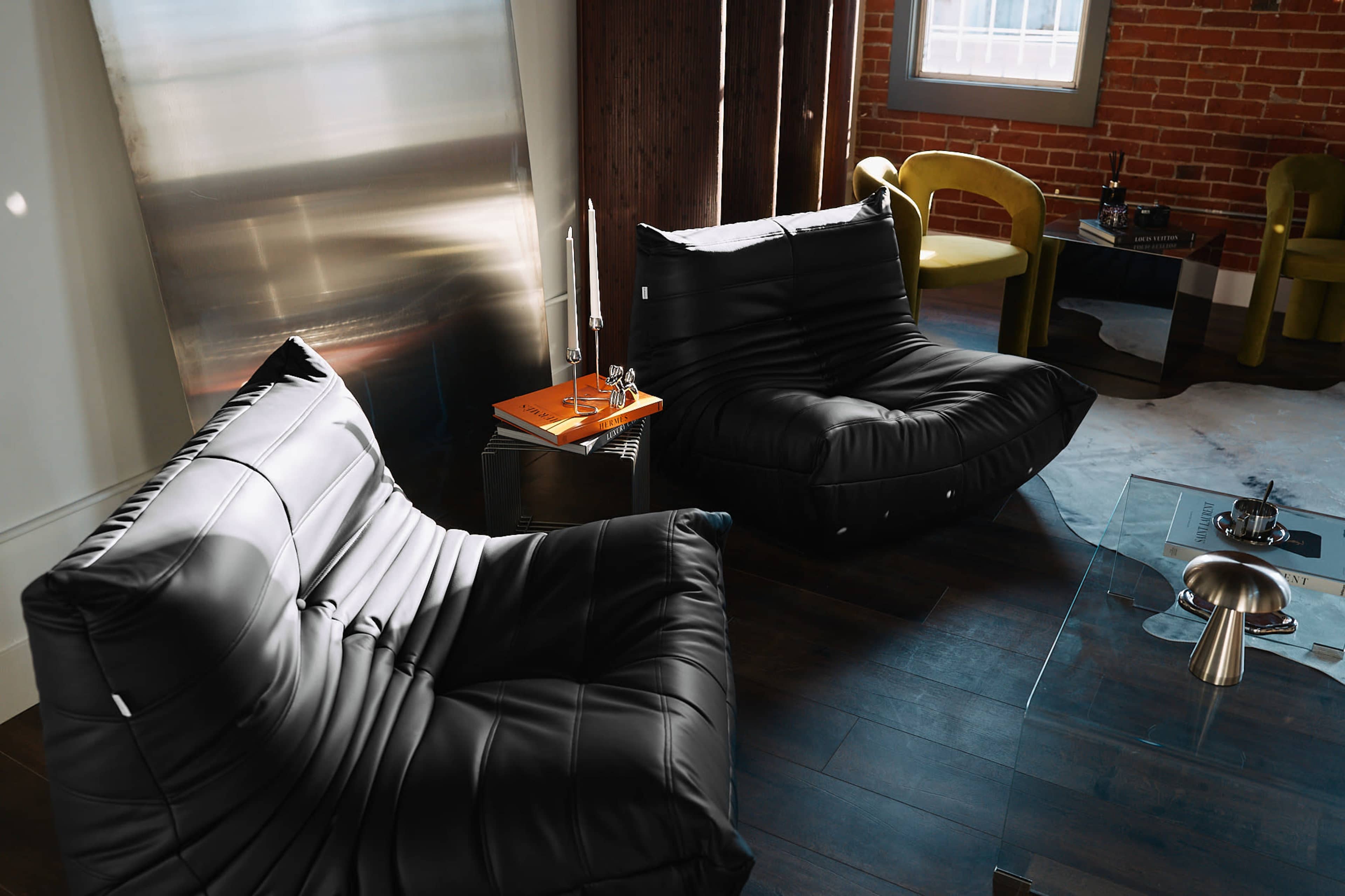 The image shows a modern interior with two large black lounge chairs, a glass coffee table, and a brick wall in the background.