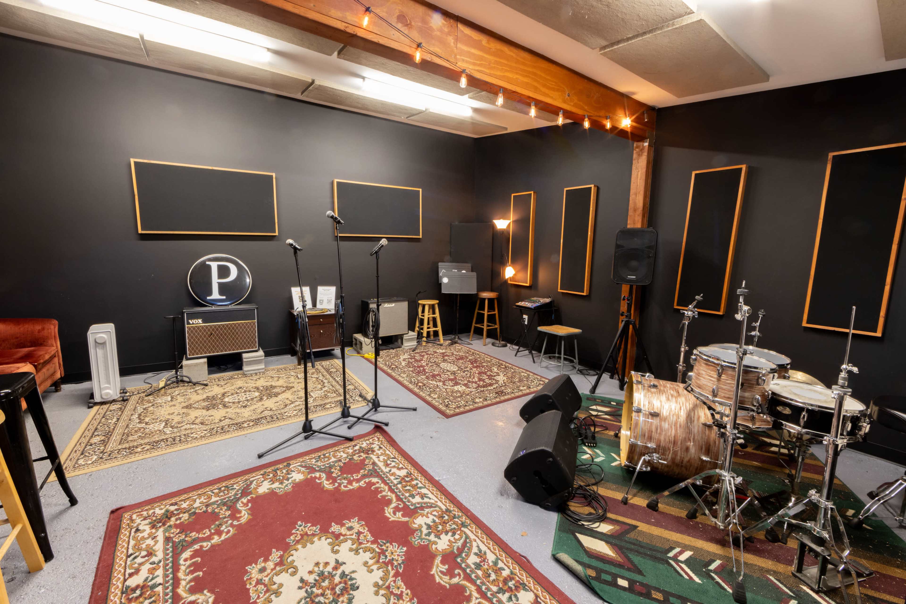 A music rehearsal space featuring two microphone stands, a drum set, amplifiers, and various seating arrangements against a backdrop of black walls and area rugs.