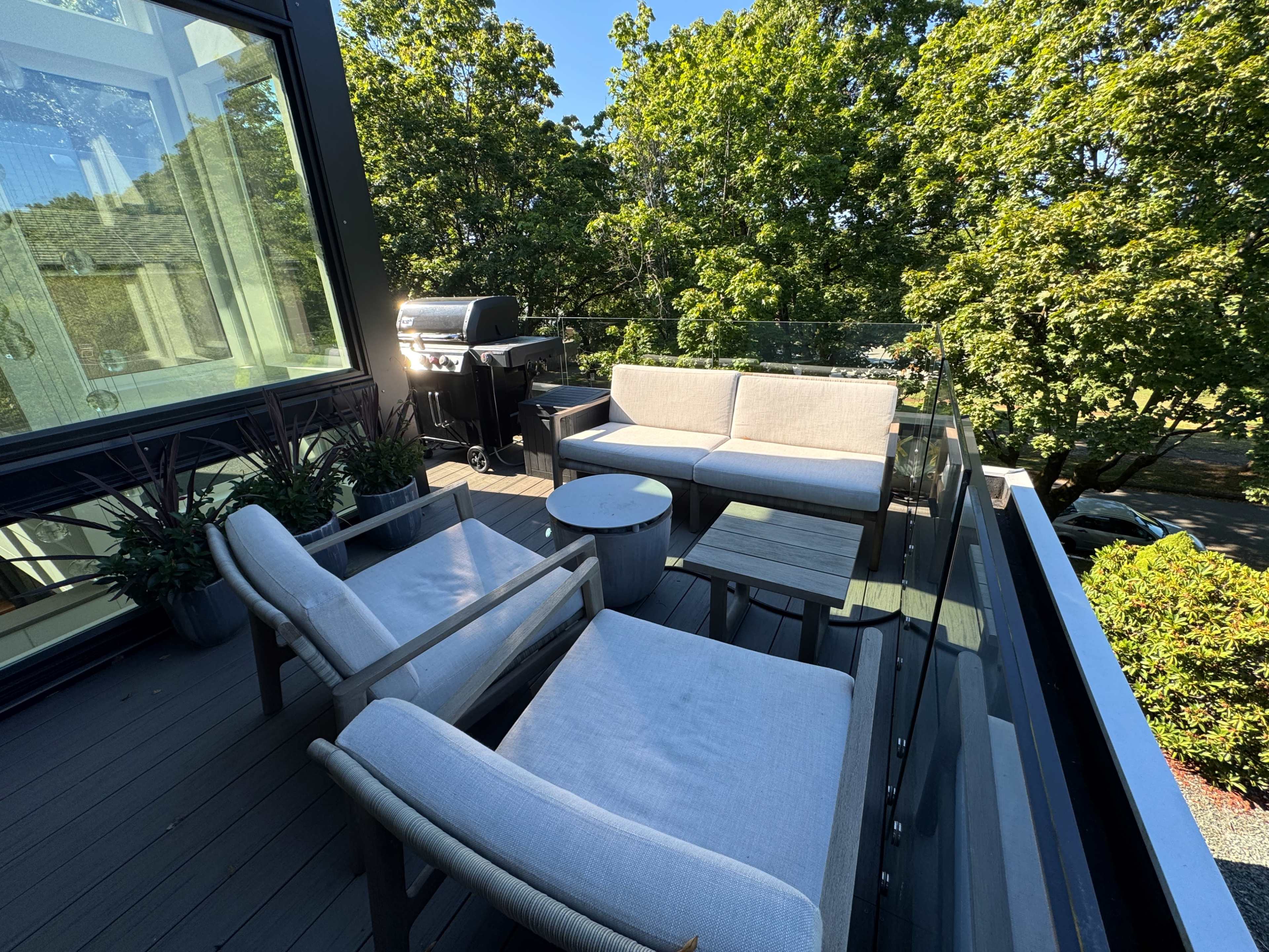 The image shows a rooftop deck furnished with two lounge chairs, a small table, and a sofa, overlooking trees and featuring a grill in the background.